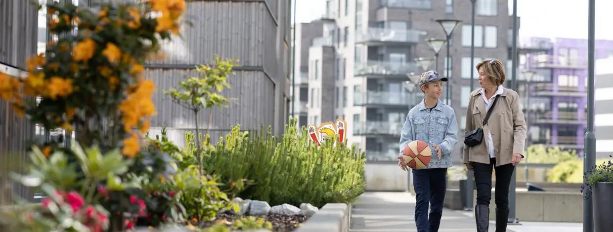 gutt og dame går utenfor blokkbebyggelse og pappfigur i bakgrunnen
