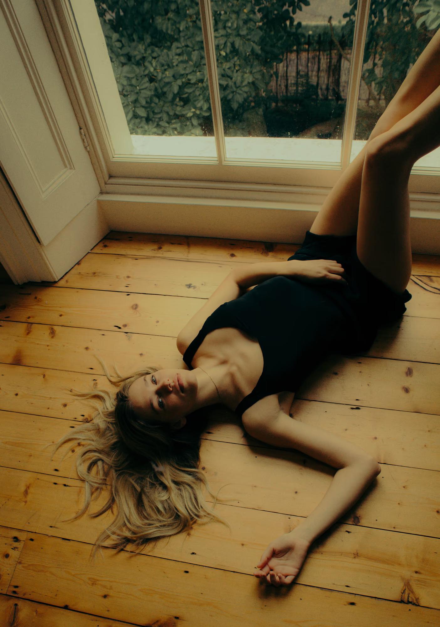 A model lies on her back on a bare wooden floor beside a tall window, wearing a black cropped top and dark shorts. One leg is raised upward, hair splayed around her head. She looks up toward the camera with a relaxed, candid expression. Soft daylight from the window fills the frame.