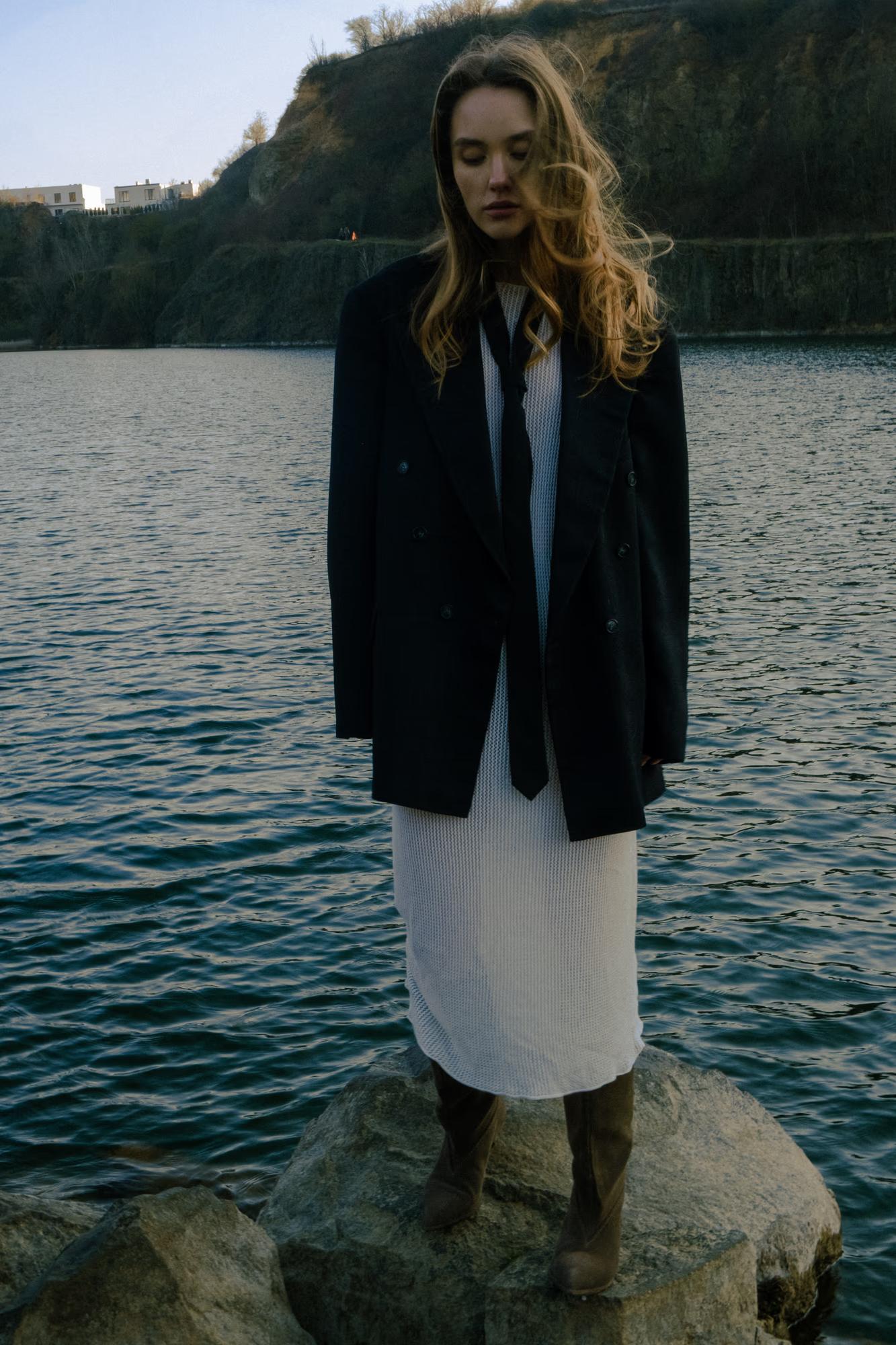 Medium full shot: The model stands on the water's edge, wearing white pants and a black blazer, facing the lake.