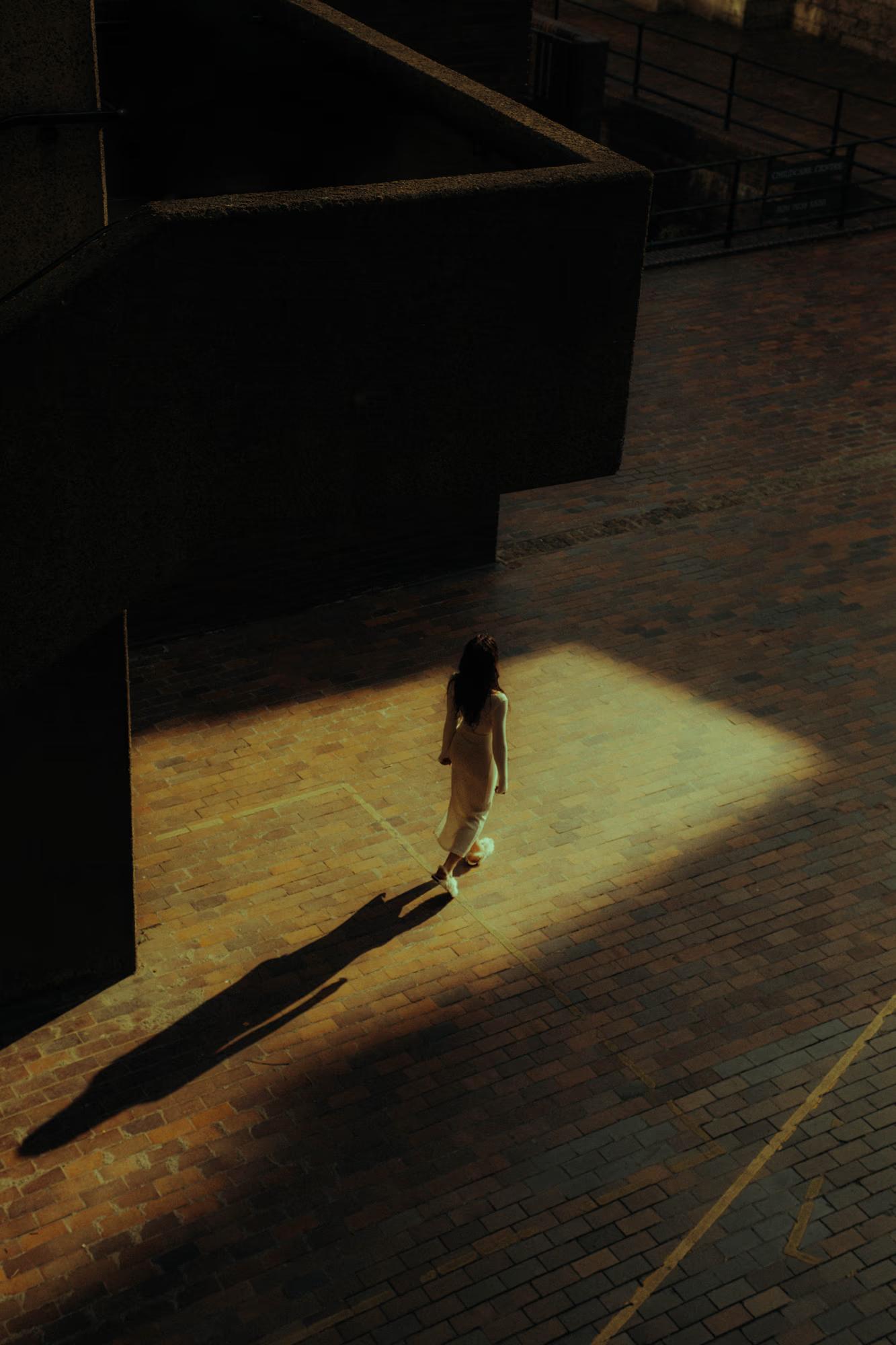 Full body fashion shot of person in dark outfit standing in dramatic shadow under London urban architecture, high contrast moody street editorial