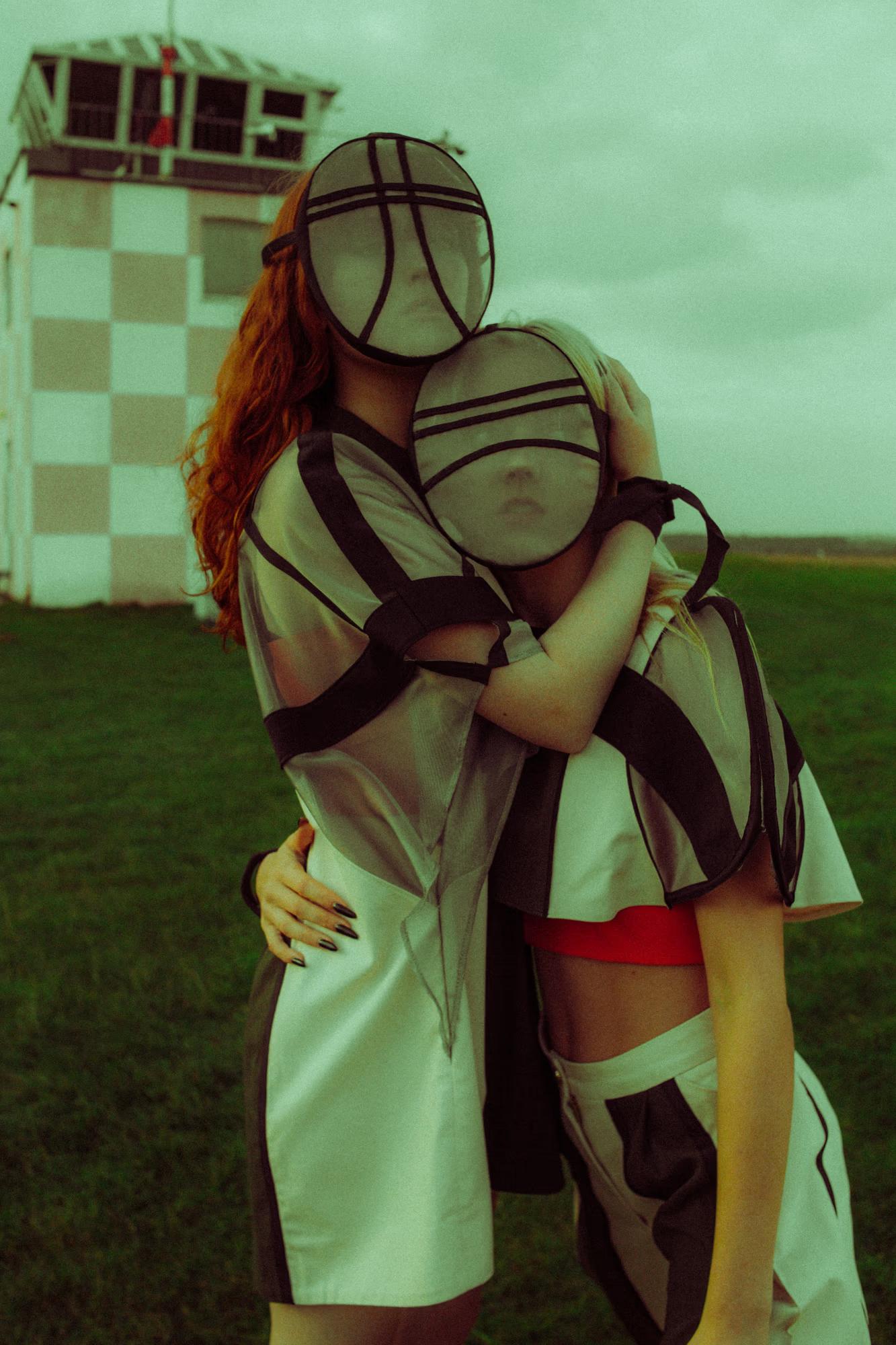 Two models embracing tightly on a grass airfield, both wearing sheer grid-patterned oval face masks that obscure their features. One has long red hair and a white structured dress; the other is blonde and wears a bold black-and-white geometric top with a red crop underneath. A checkered aviation control tower is visible in the background.