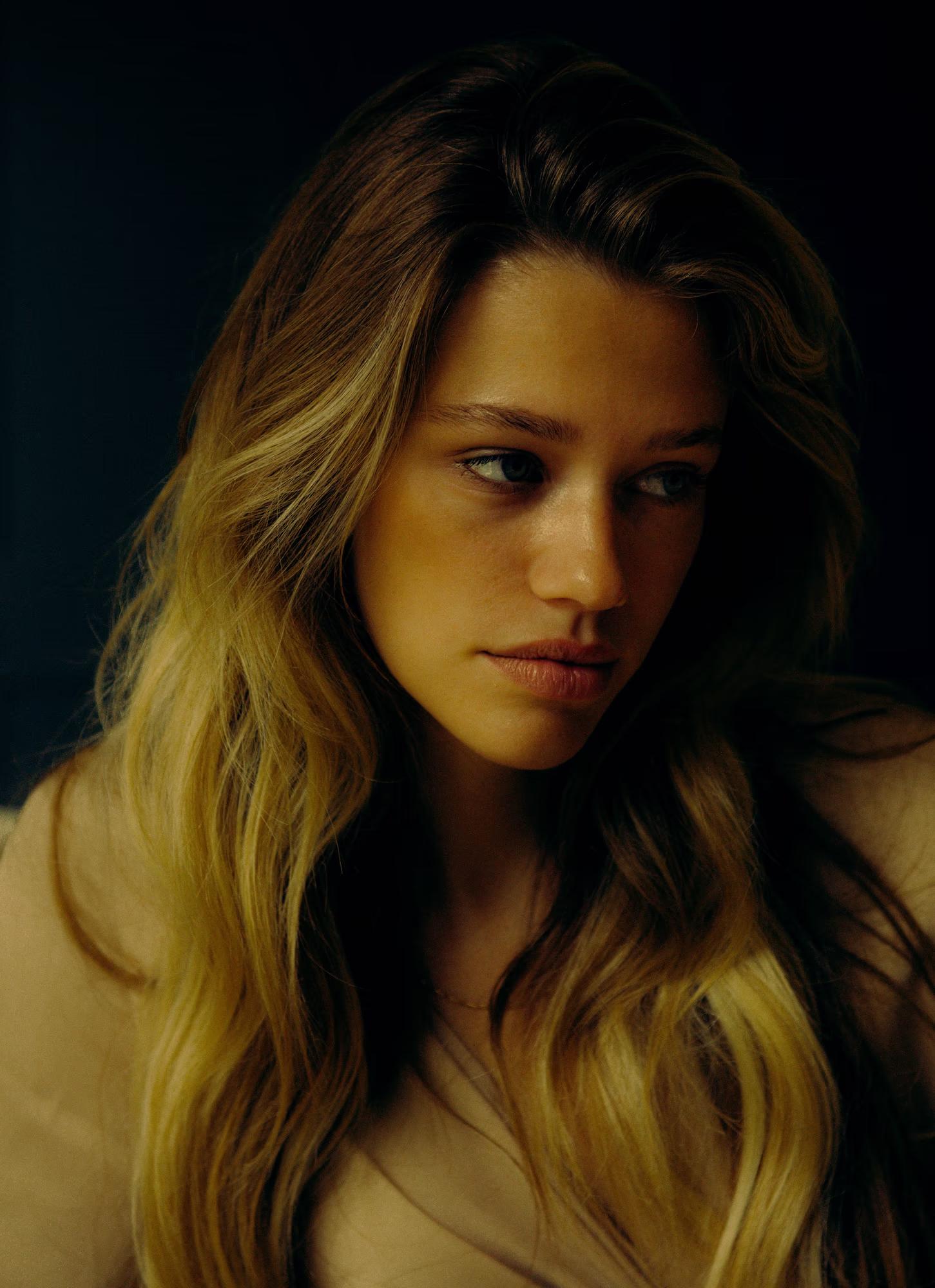 Close-up portrait of a model with long wavy blonde hair, gazing at the camera with a calm, direct expression. She wears a soft nude-toned top. Warm amber light illuminates her face against a near-black background, highlighting her blue-green eyes and natural freckles.