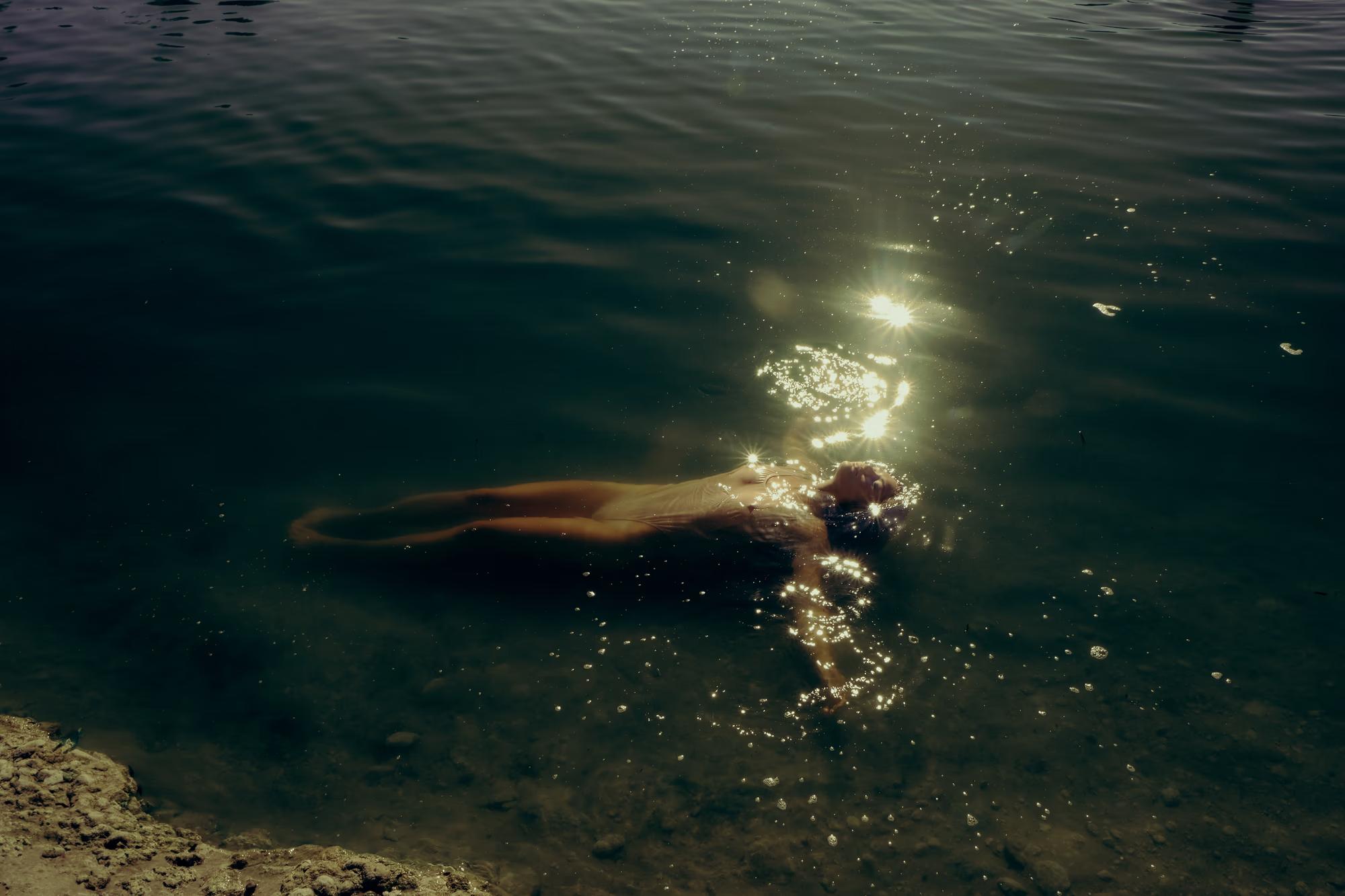 Underwater wide shot of woman swimming in dark teal lake water with sunlight rays piercing through surface, cinematic fine art photography