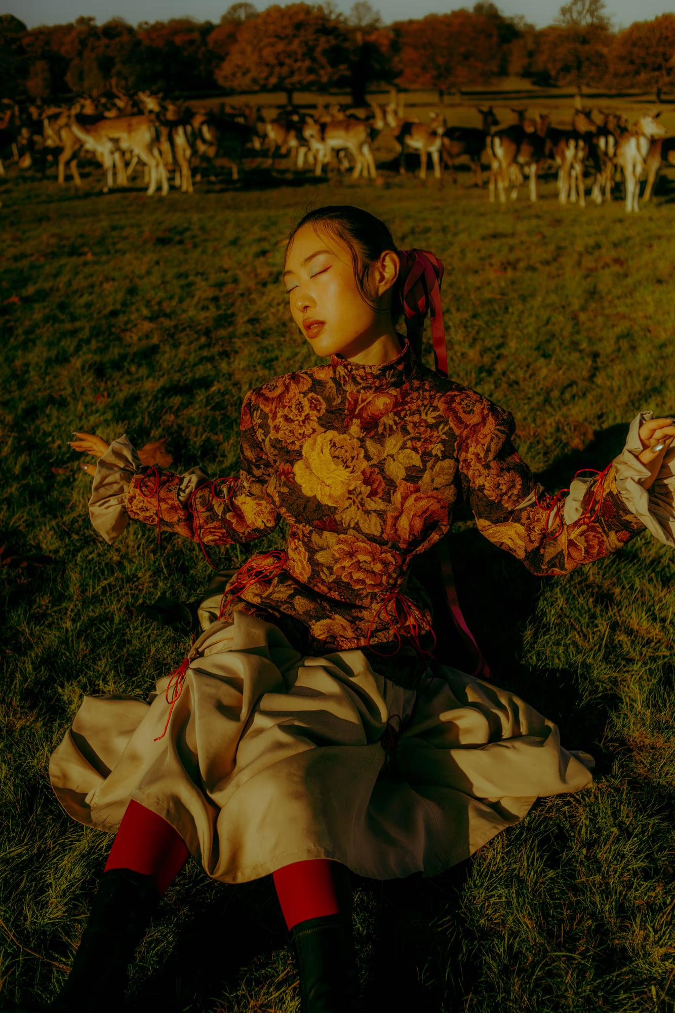 Medium fashion shot of woman in bold autumn-toned floral designer dress among trees, warm cinematic light, Richmond Park London luxury editorial