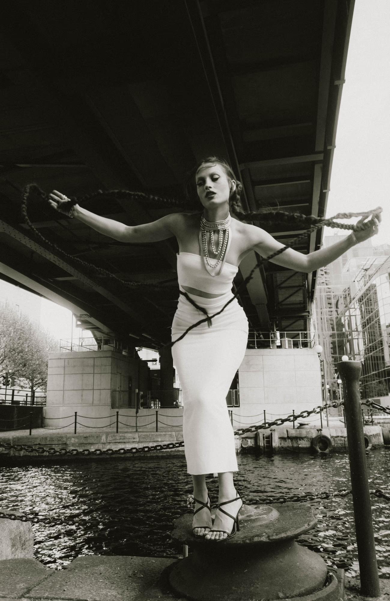 Full body shot of female model in light dress posing with arms raised, black and white editorial, Canary Wharf London