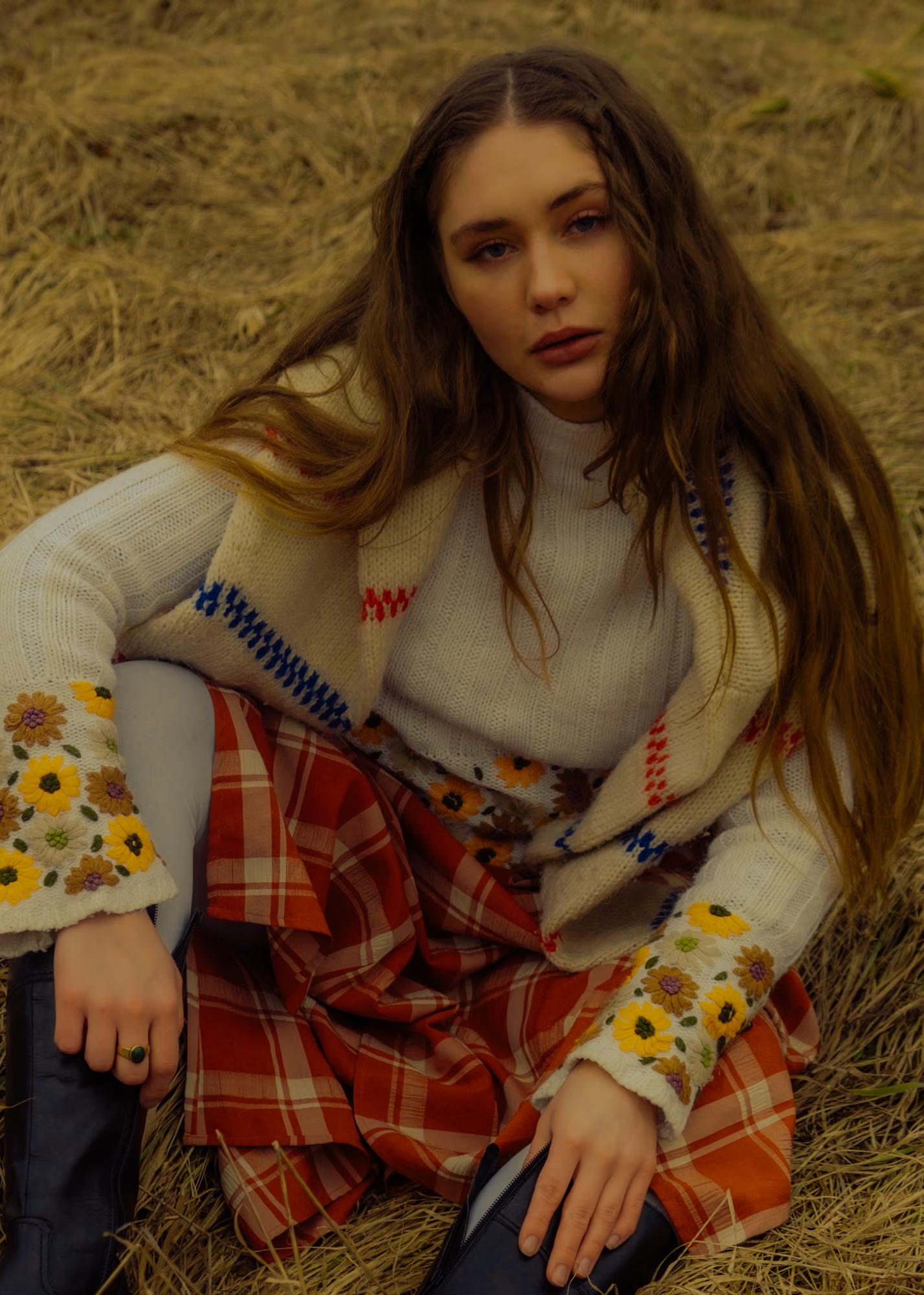 Medium close-up fashion shot of woman with long dark hair in white blouse seated in dry Carpathian grass, warm golden tones, ethno style portrait