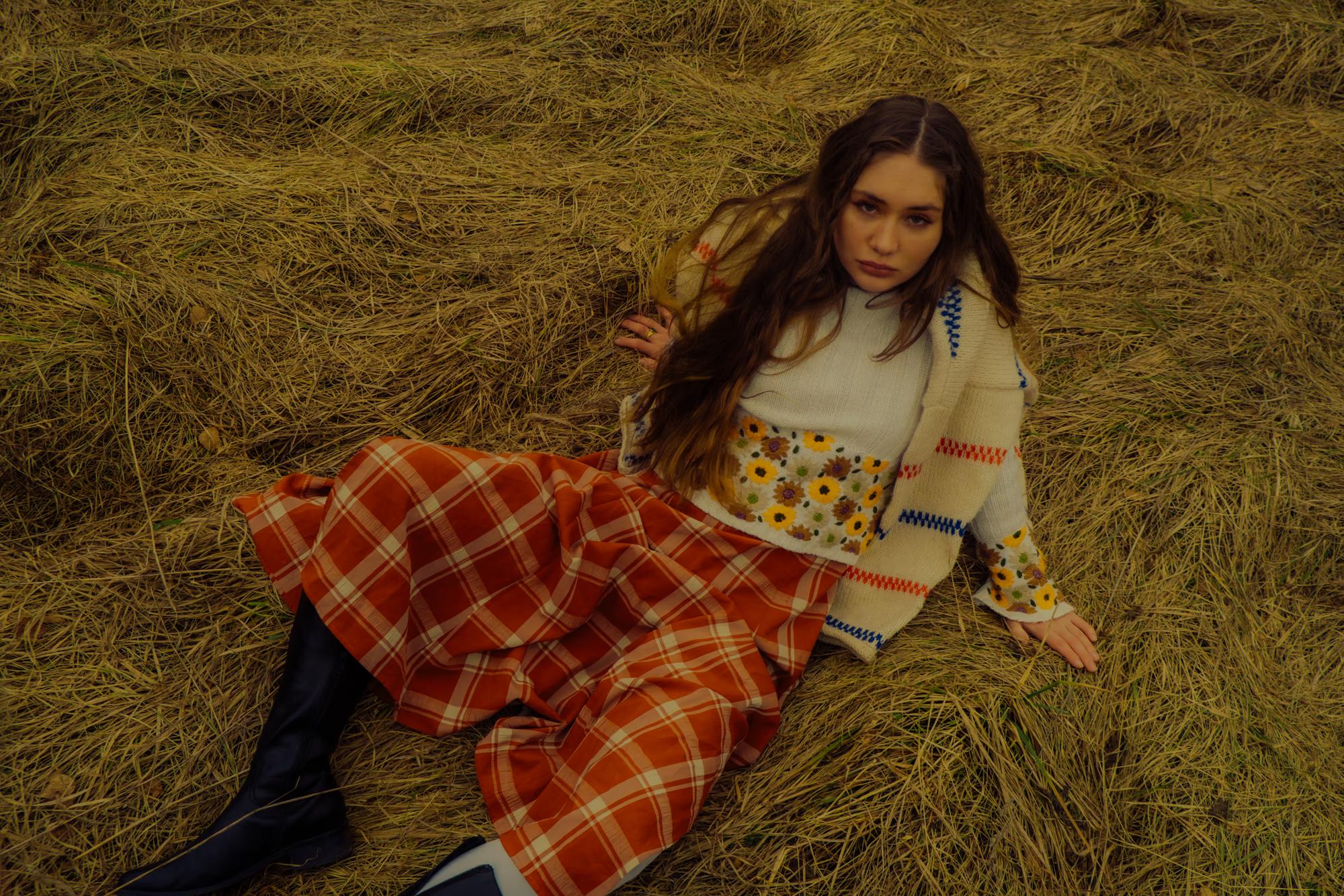 Full body fashion shot of woman in white blouse and bold orange-red tartan maxi skirt sitting in dry golden grass, warm autumn tones, Carpathian ethno editorial