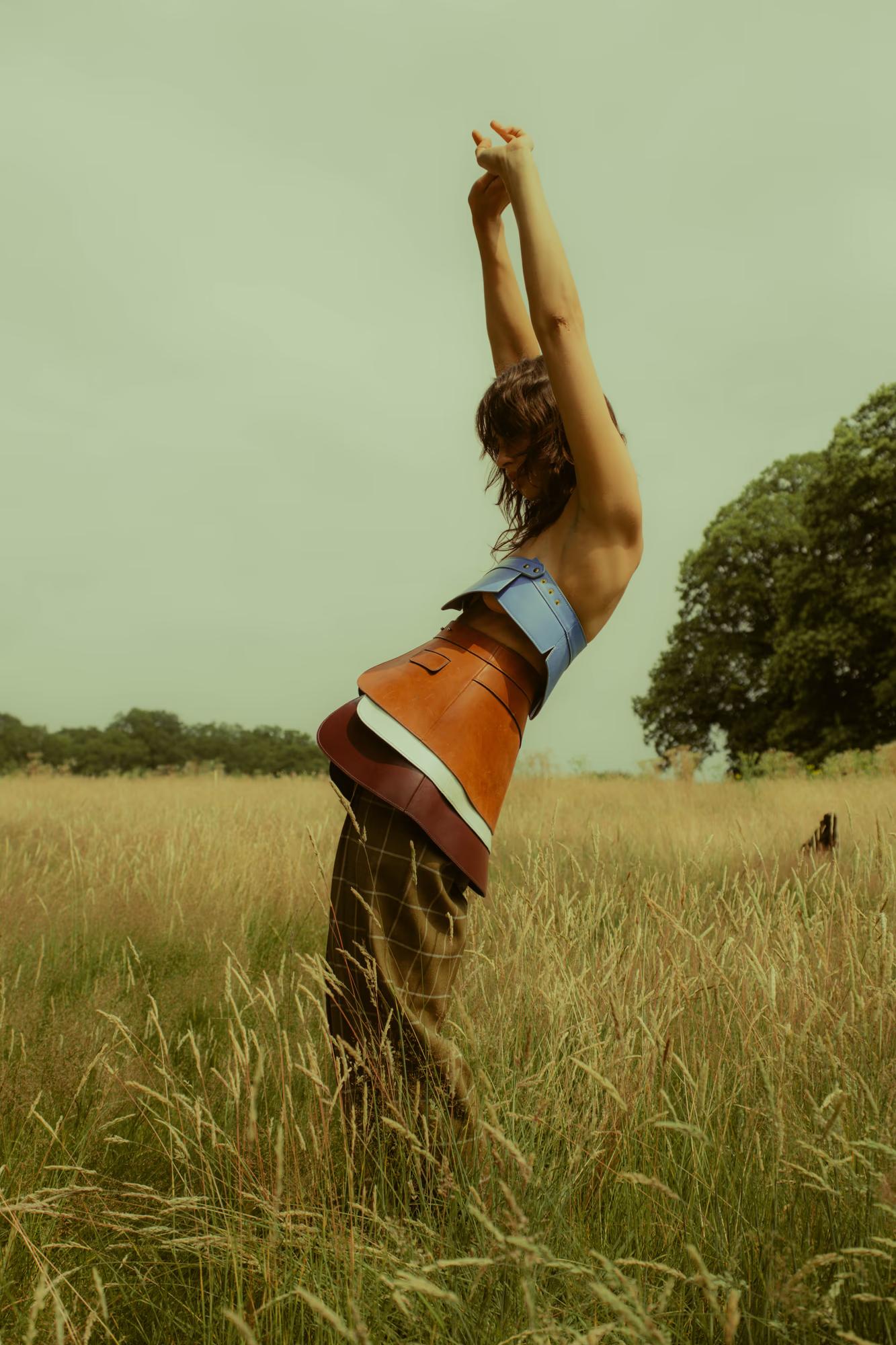 A model stands sideways in a tall golden grass field, arms raised straight above her head. She wears a structured colourblock corset-style piece in sky blue and tan leather tones, layered over wide-leg dark trousers. The pale teal sky and a distant oak tree frame the dynamic, sculptural pose.
