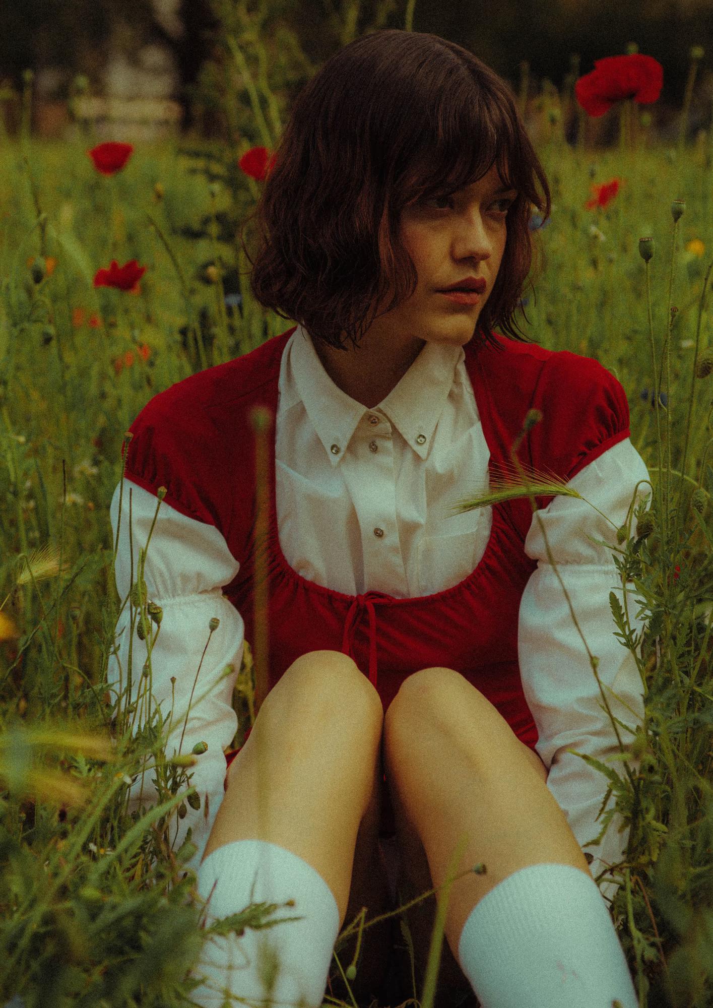  A model sits in the middle of a wildflower meadow filled with red poppies and green stems. She wears a deep red puff-sleeved mini dress layered over a white collared long-sleeve shirt, with white knee-high socks. Her dark bob falls loosely around her face as she gazes to the side.