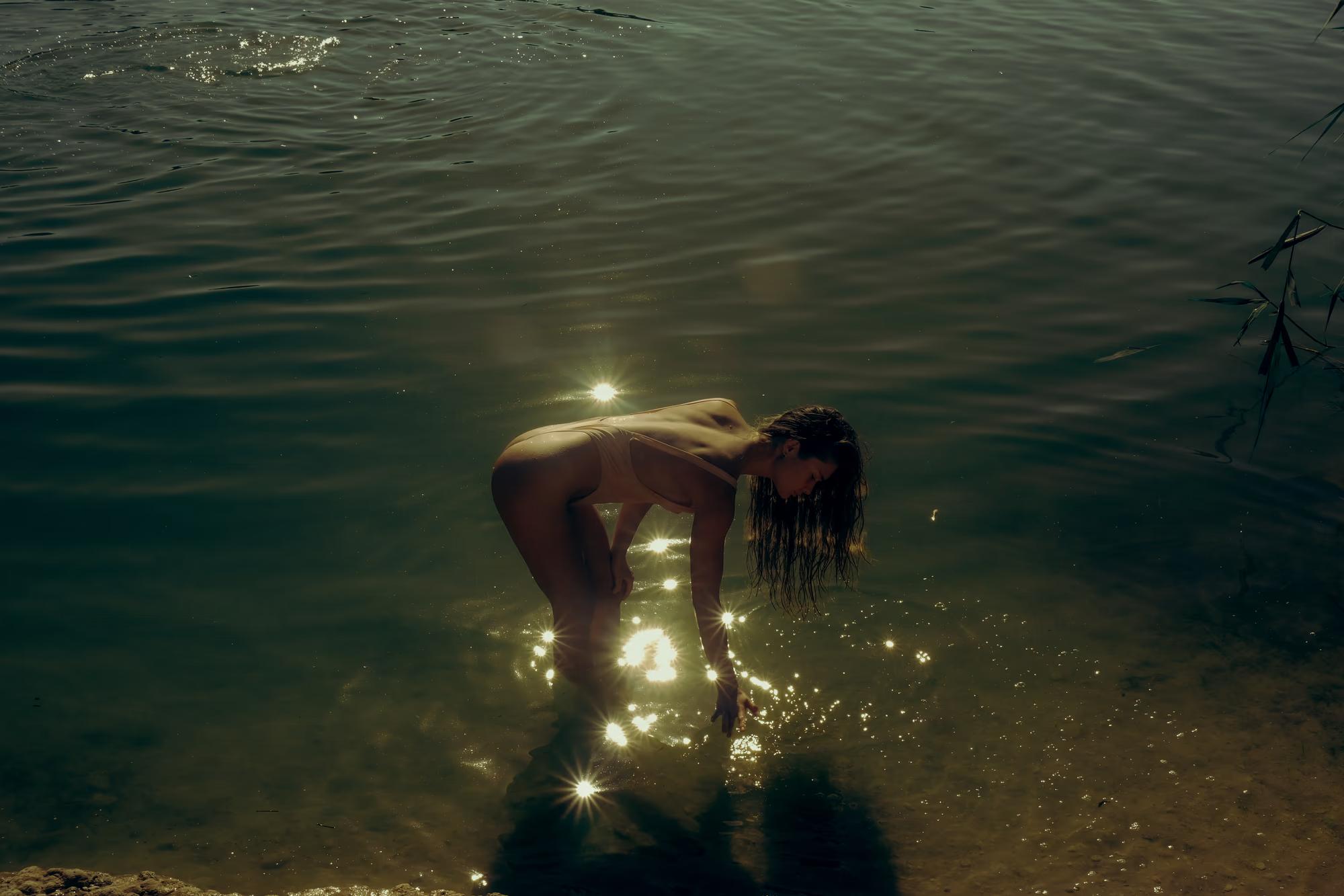 Underwater wide shot of woman floating in dark lake with warm light source visible, mysterious cinematic atmosphere, fine art photography