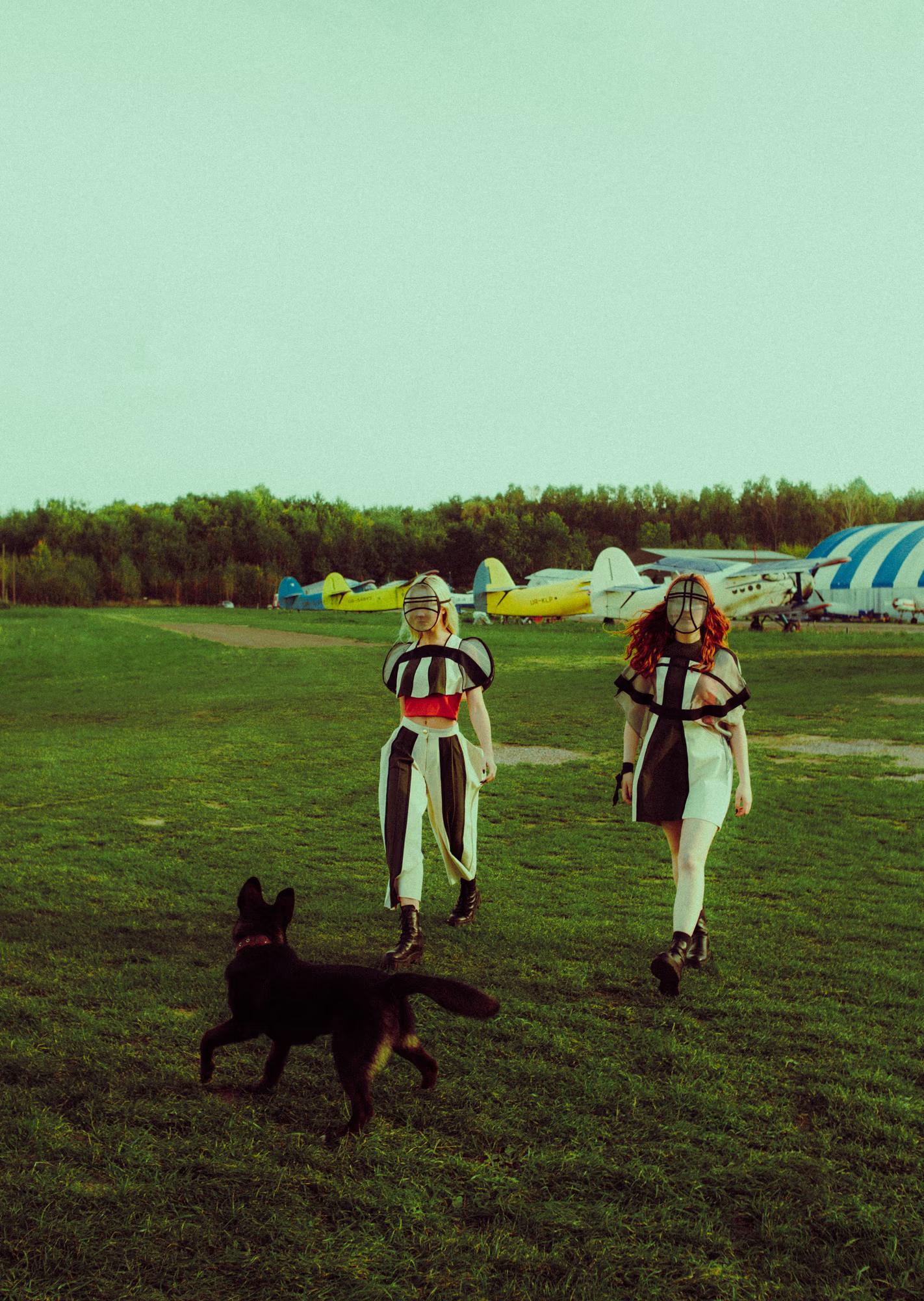 Two models in geometric black-and-white outfits and sheer oval face masks walk across a broad grass airfield. A black dog trots in the foreground. In the background, a row of colourful small planes — yellow, white, and blue — lines the far edge of the field against a treeline.