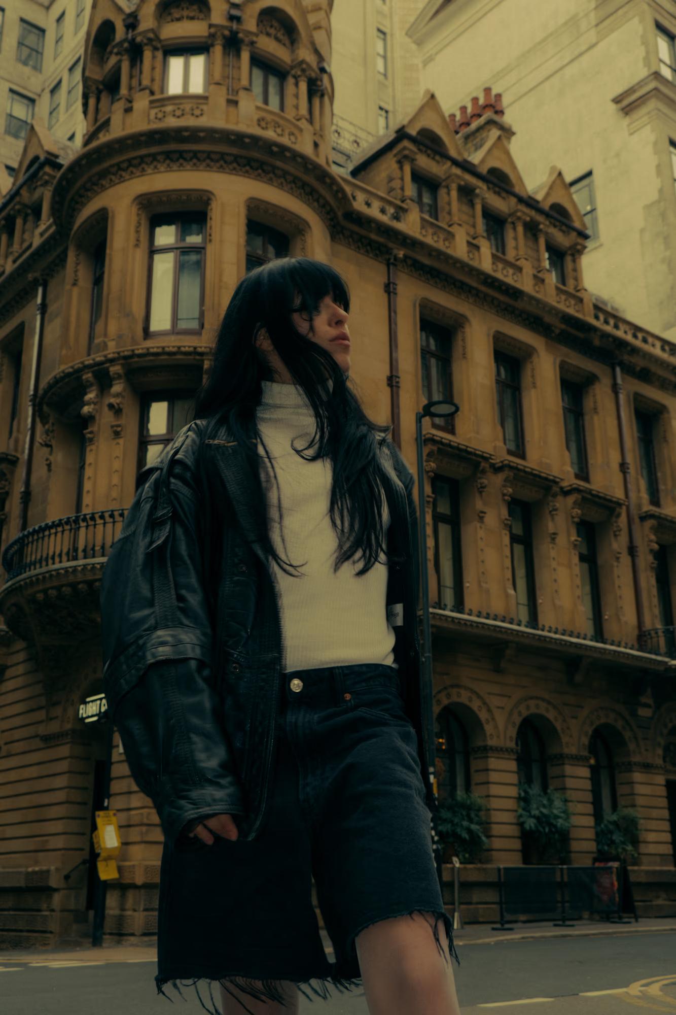 Full body fashion shot of person in dark coat and trousers standing on Manchester street with classic Victorian architecture in background, moody urban editorial