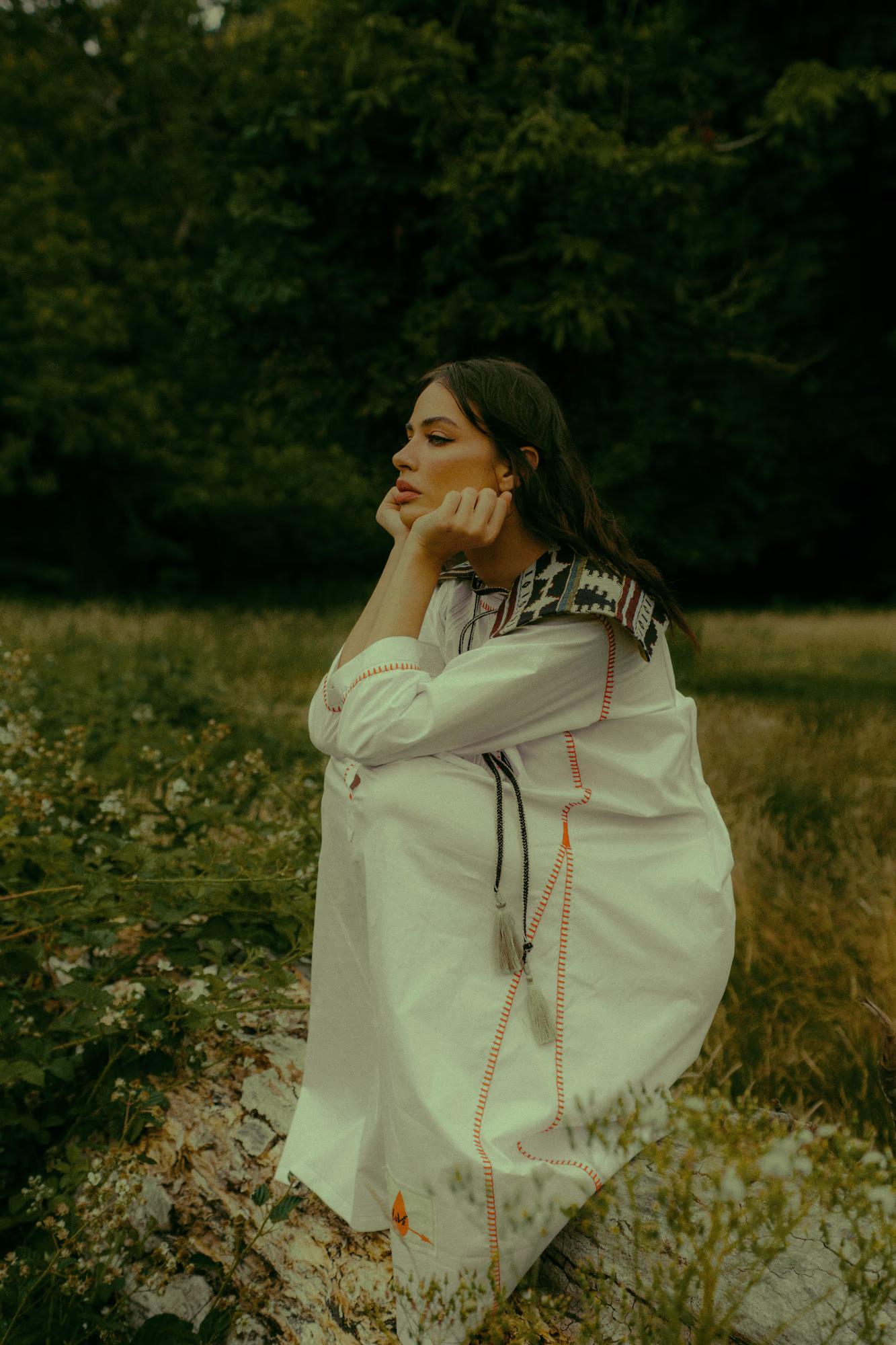 A model sits on a fallen log in a park, resting her chin on her hand in a thoughtful pose. She wears a white long-sleeved folk dress with orange running stitch borders, a geometric woven yoke, and a long tassel cord tie. She looks to the side against a dark backdrop of trees and low greenery.
