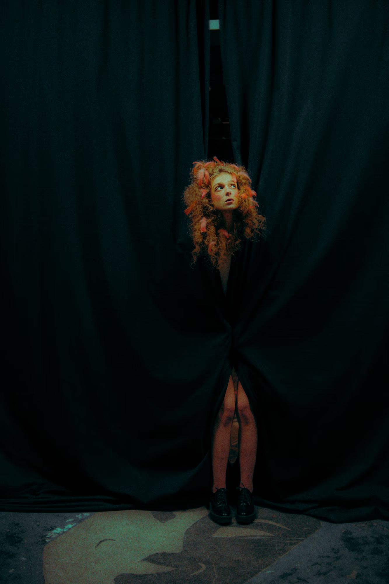 Minimalist fashion shot of a model standing between heavy velvet stage curtains in the dark.