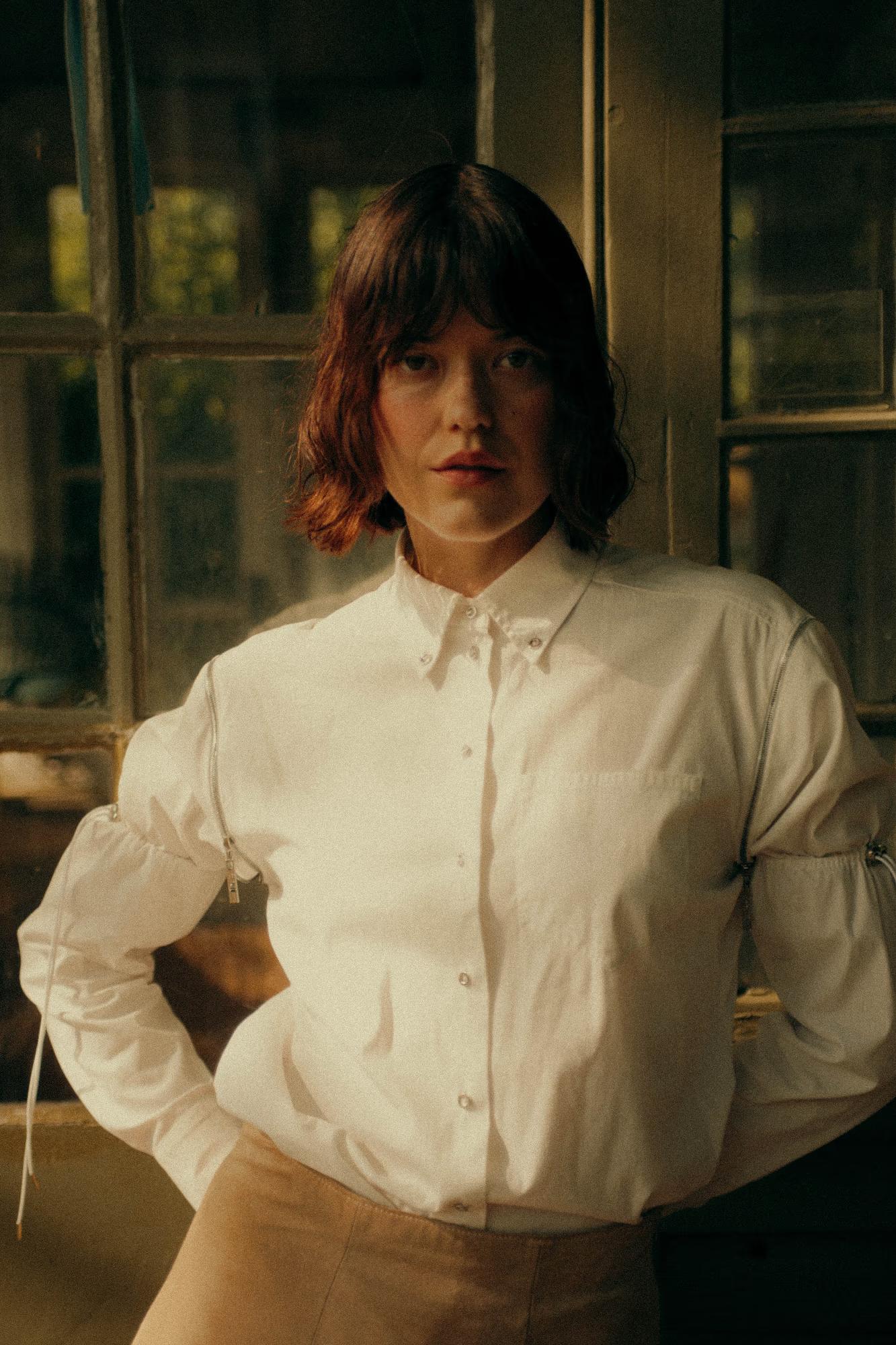 Three-quarter portrait of a model standing before old greenhouse windows, hands behind her back. She wears an oversized white cotton shirt with distinctive zip detailing along the sleeves and a sand-coloured skirt visible at the hem. Her dark bob and direct gaze give the image a cool, composed mood.