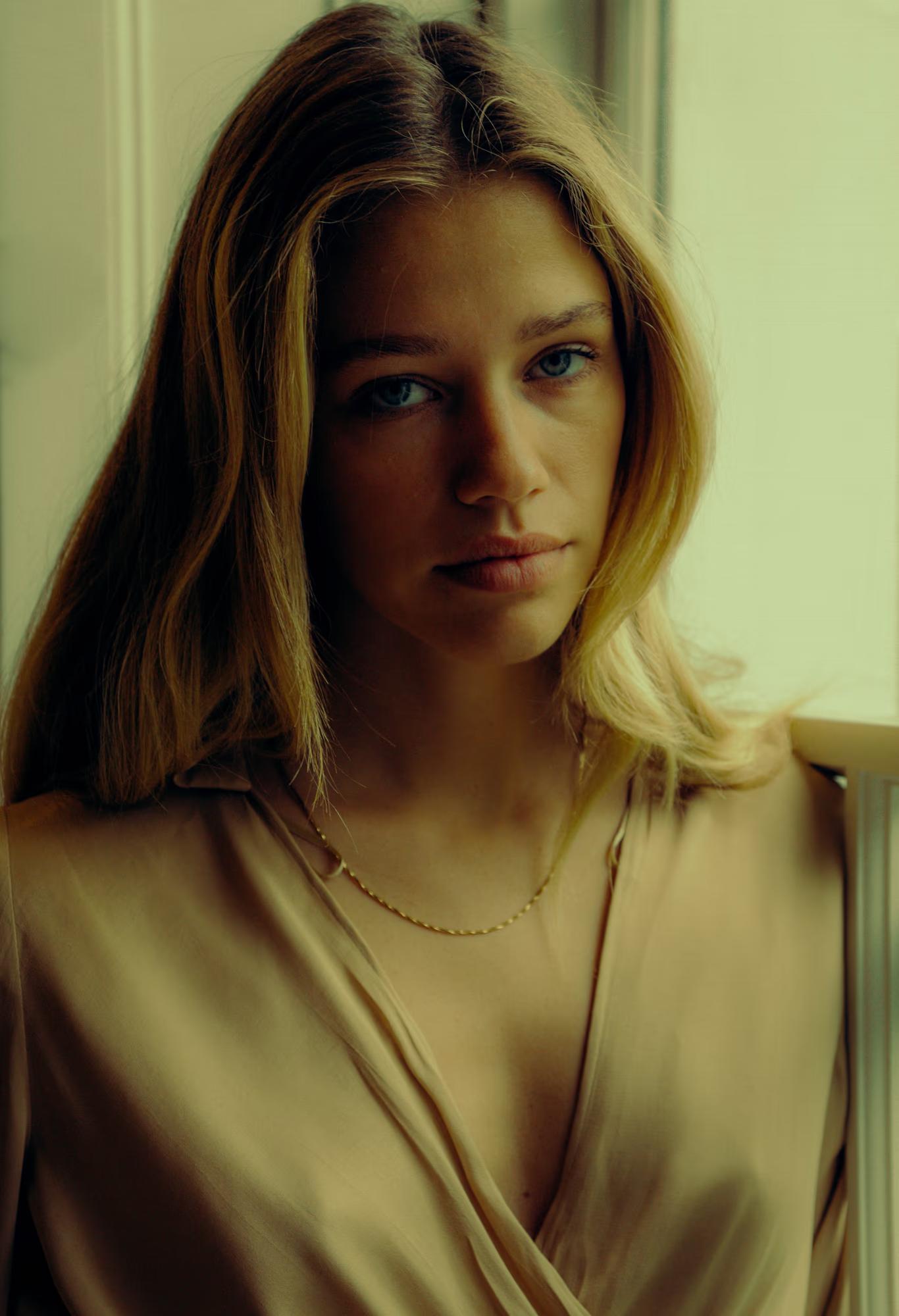 Close-up portrait of a model with straight blonde-highlighted hair and pale blue eyes, wearing a draped satin wrap in a warm sand tone and a delicate gold link necklace. She faces the camera with a quiet, steady gaze. Soft window light brightens the left side of her face.