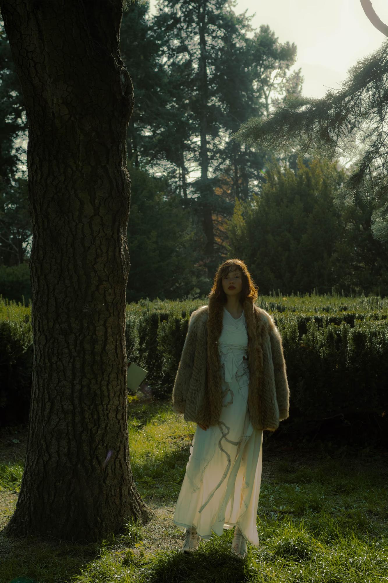 fashion art shot of woman in light outfit standing in dark forest with dramatic backlight through trees, ethereal dark fantasy editorial