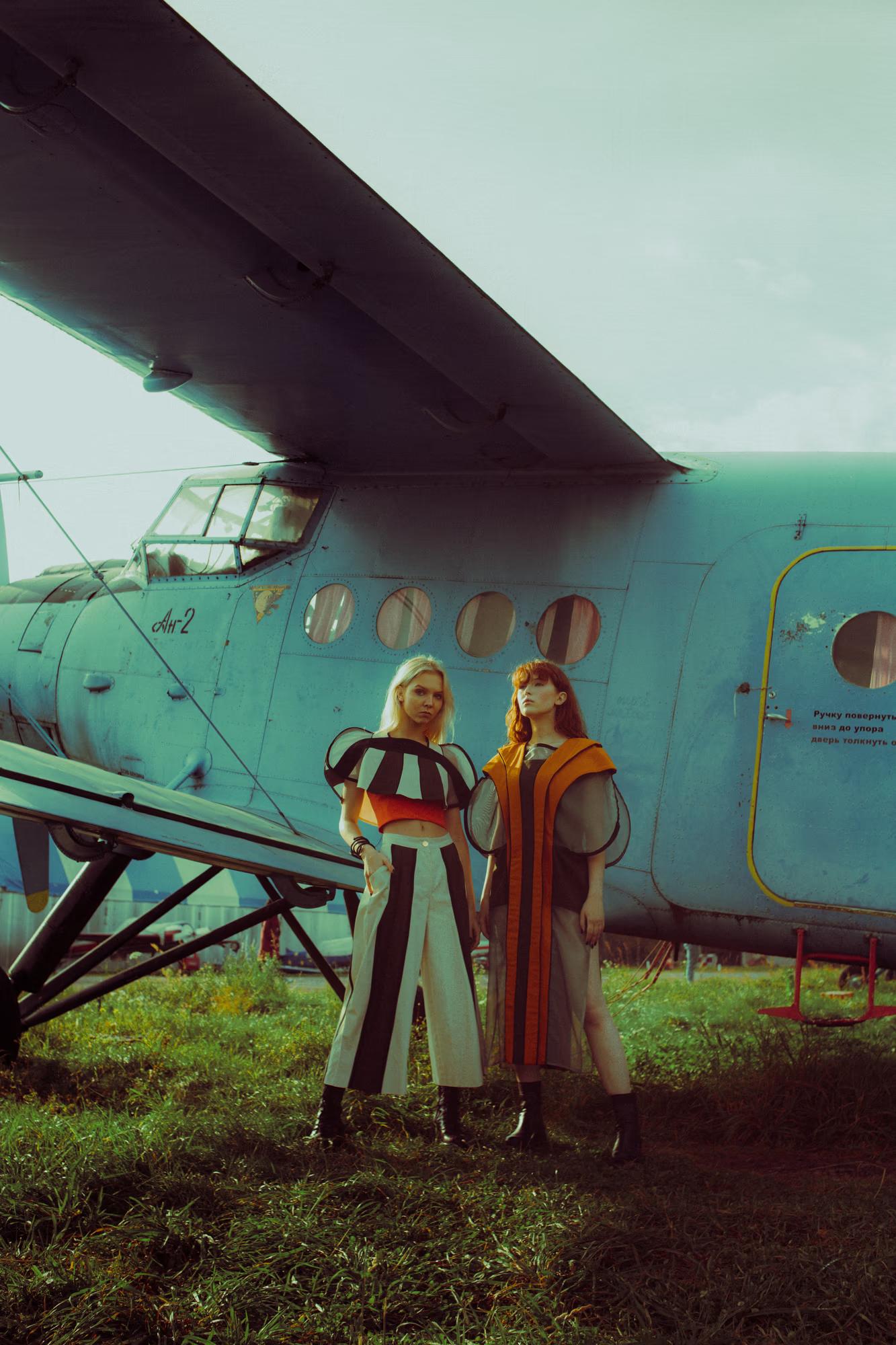 Two models standing side by side on grass in front of a weathered turquoise Antonov An-2 biplane. The blonde model wears a bold black-and-white striped wide-leg ensemble with a sculptural puffed top; the redheaded model wears an amber-striped sheer coat over a dark dress. Both wear black ankle boots.