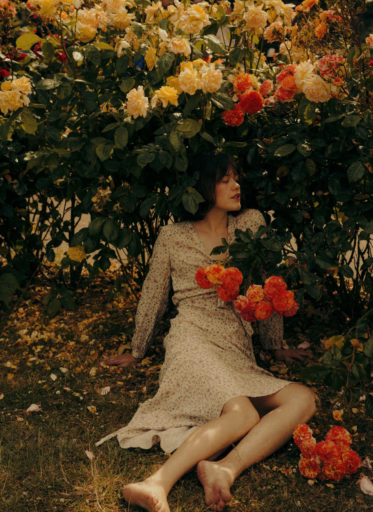 A barefoot model sits on the grass at the base of a lush rose bush bursting with orange and yellow blooms. She wears a delicate long-sleeve floral midi dress and holds a cluster of orange roses against her body, eyes closed and head tilted back.