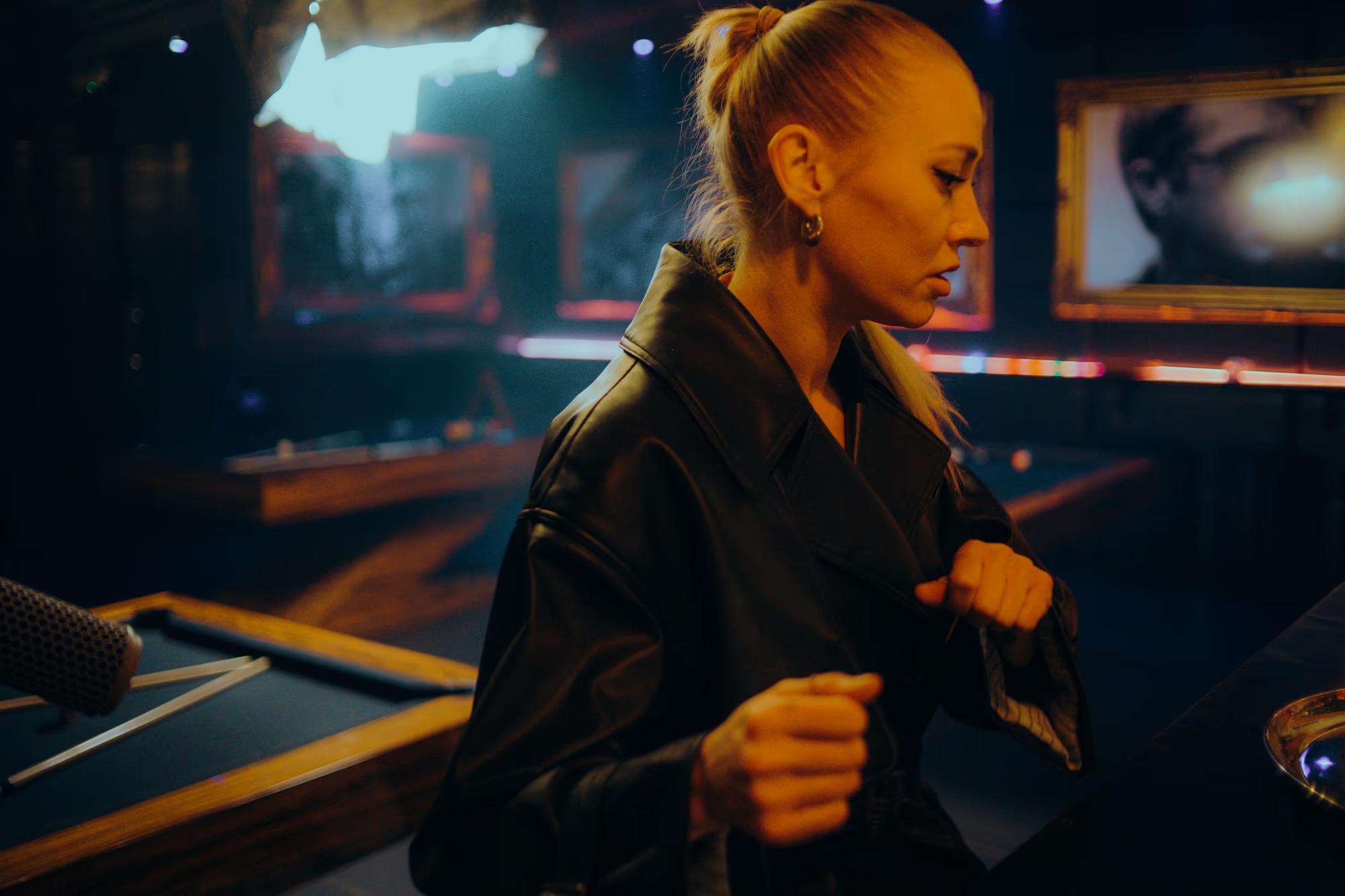 An actress in a leather jacket posing in the neon-lit interior of a London club film set.