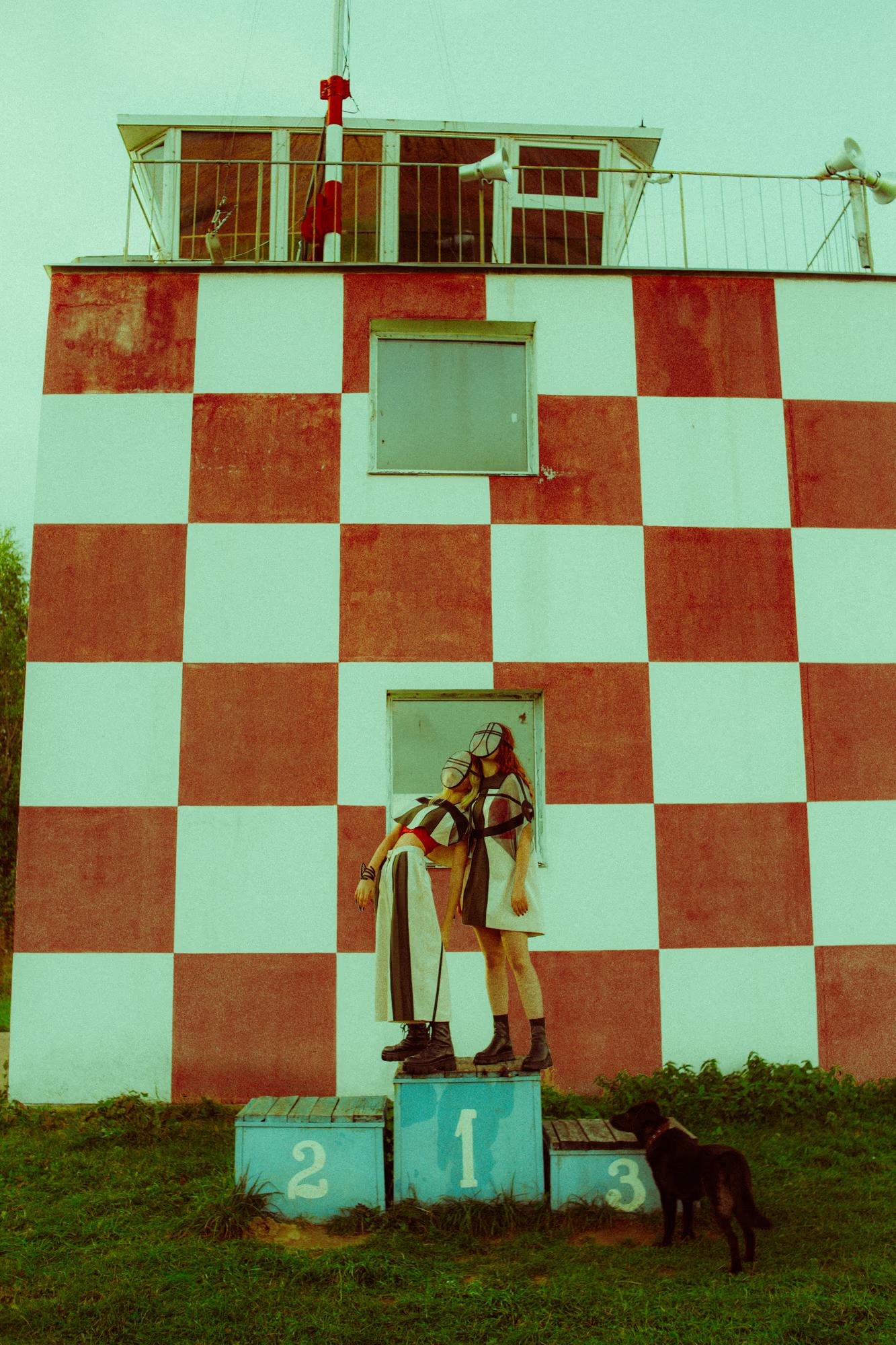 A single model wearing a sheer grid face mask and a layered geometric outfit stands atop the first-place block of a vintage podium numbered 1, 2, 3, set against the red-and-white checkered wall of an airfield control tower. A black dog stands beside the podium. The pose is theatrical and still.