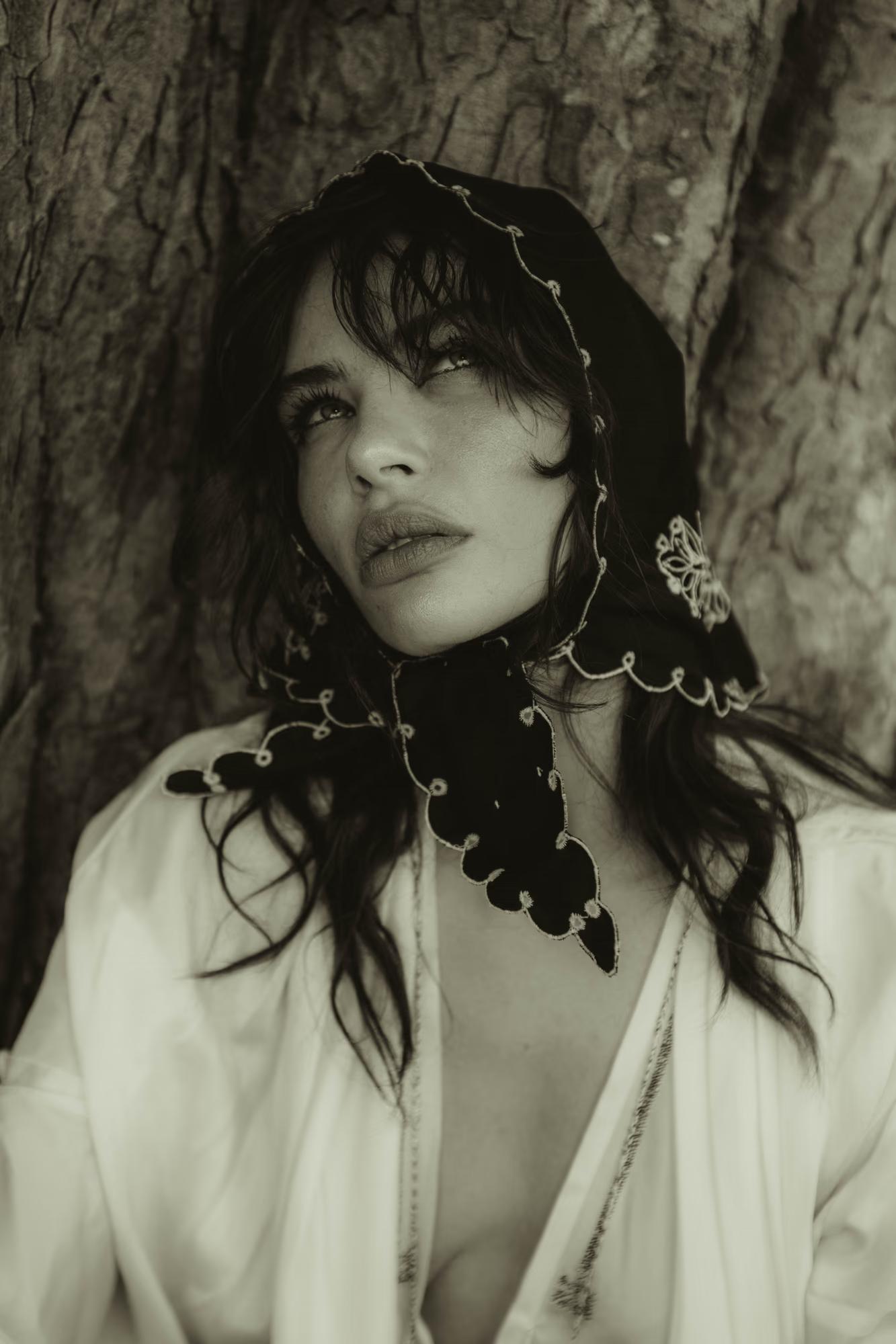 Black-and-white close-up portrait of a model leaning against the rough bark of a large tree. She wears a dark embroidered headscarf with white scalloped trim and floral details, tied loosely around her head and neck, paired with an open white dress. She looks upward with a slightly parted expression, dark wavy hair escaping the scarf.