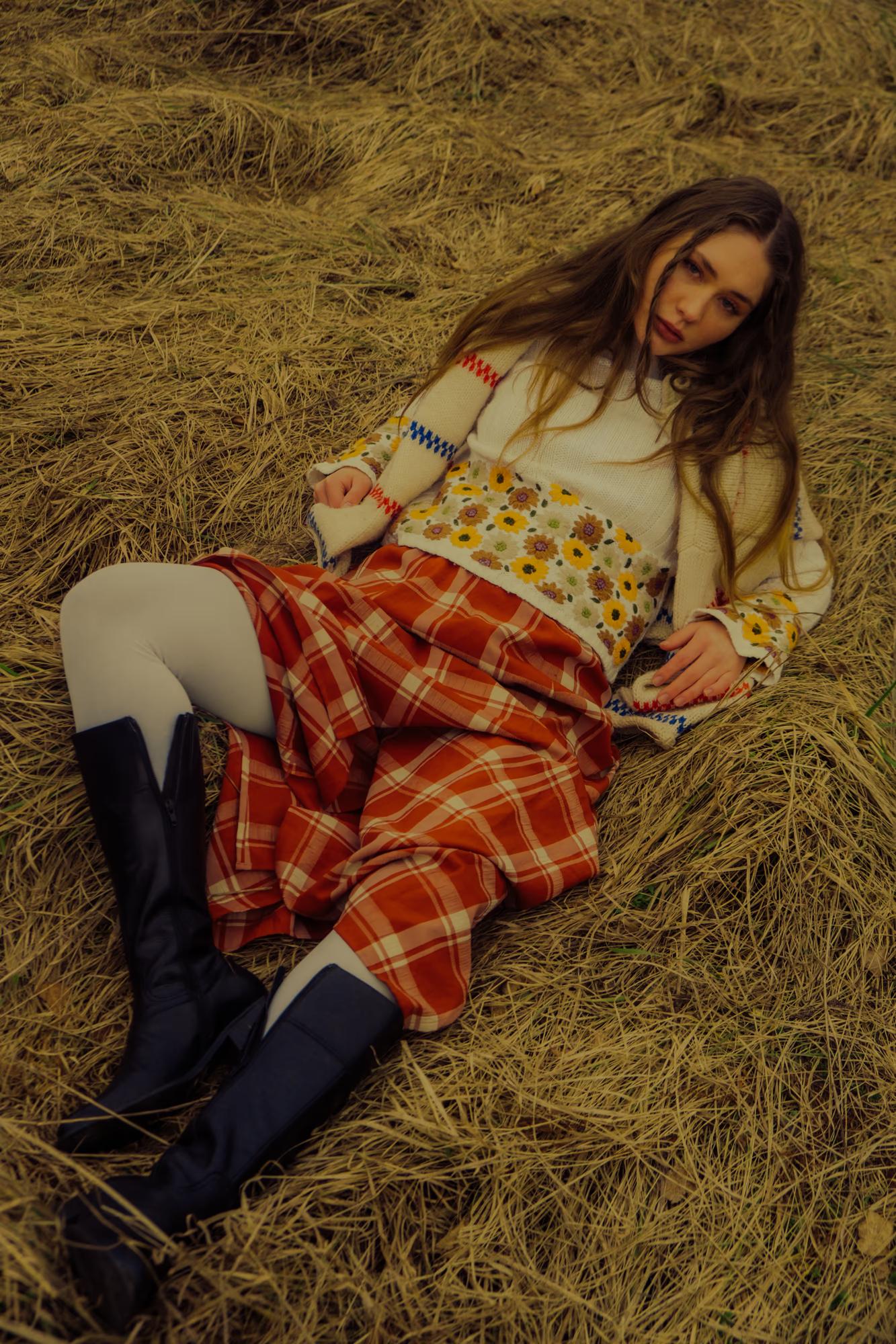 Medium fashion shot of woman in floral and tartan outfit crouching in golden Carpathian meadow, vibrant autumn colors, ethno fashion editorial
