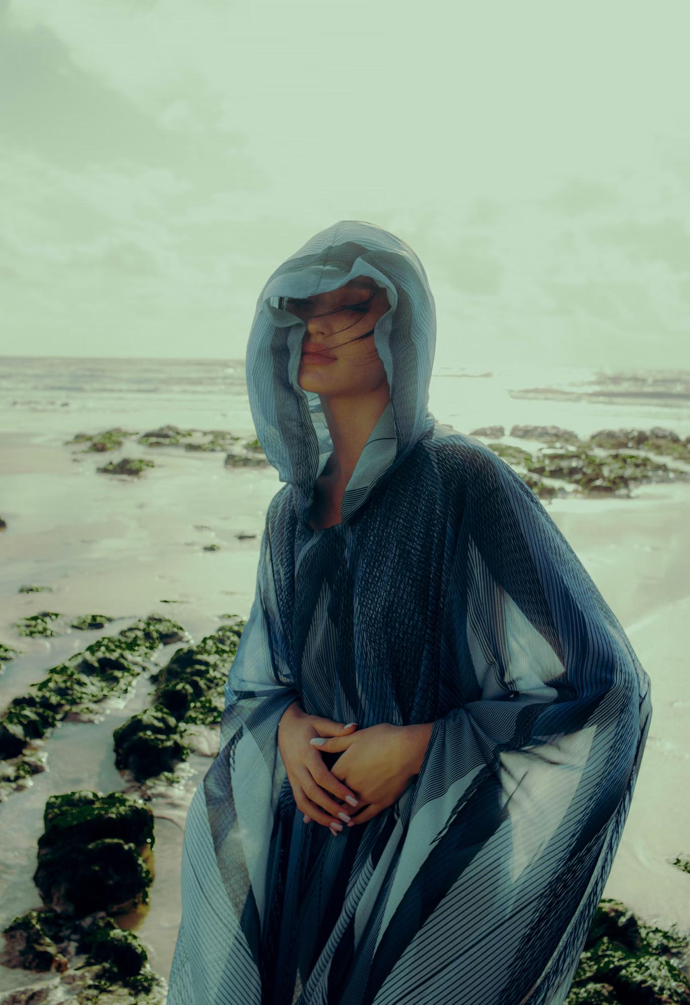 Medium fashion campaign shot of woman in blue designer cape seated on Eastbourne coastal rocks, moody overcast atmosphere, luxury editorial