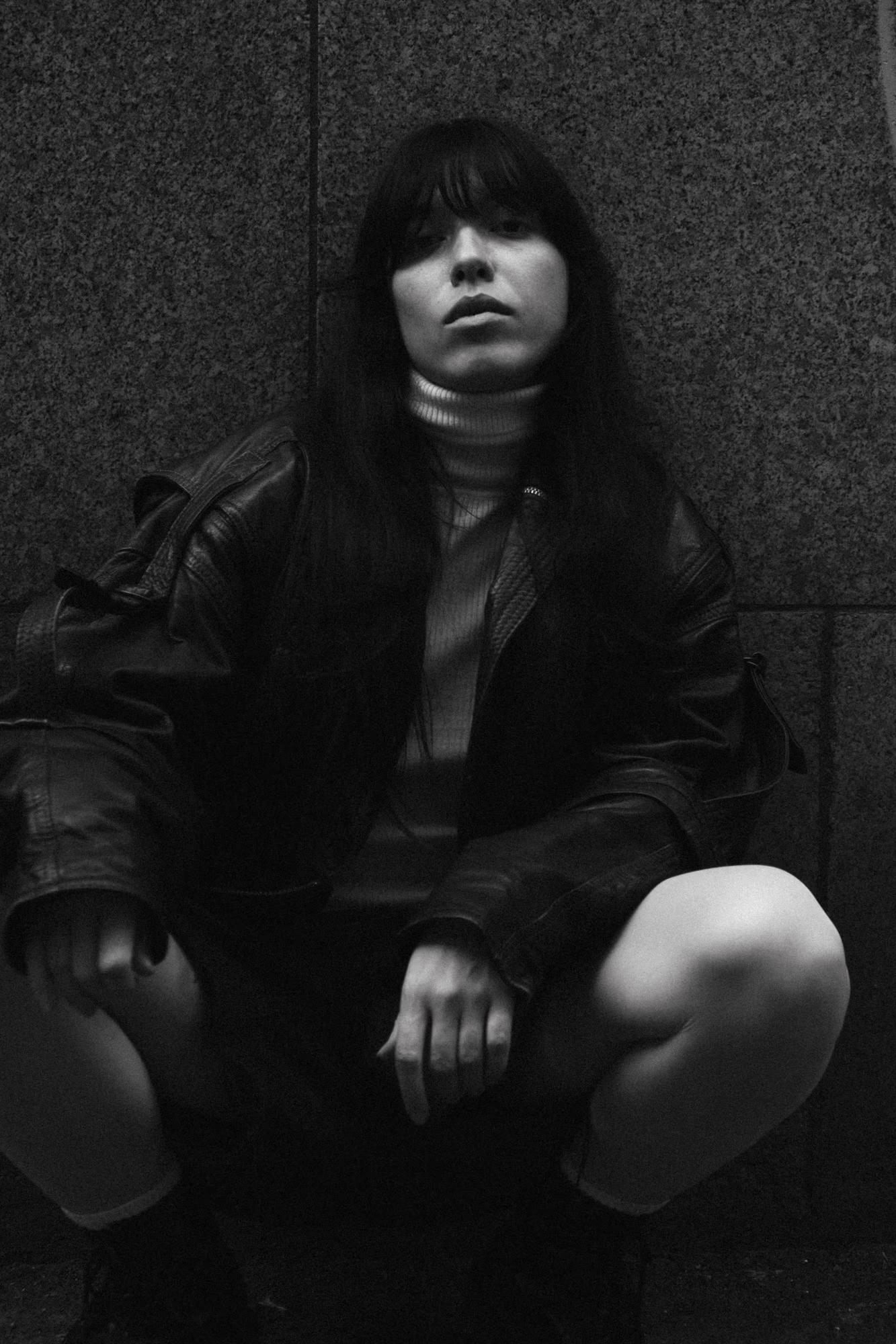 Full body dark BW fashion art shot of person in all black outfit seated on ground, dramatic low key lighting, moody Manchester urban editoria