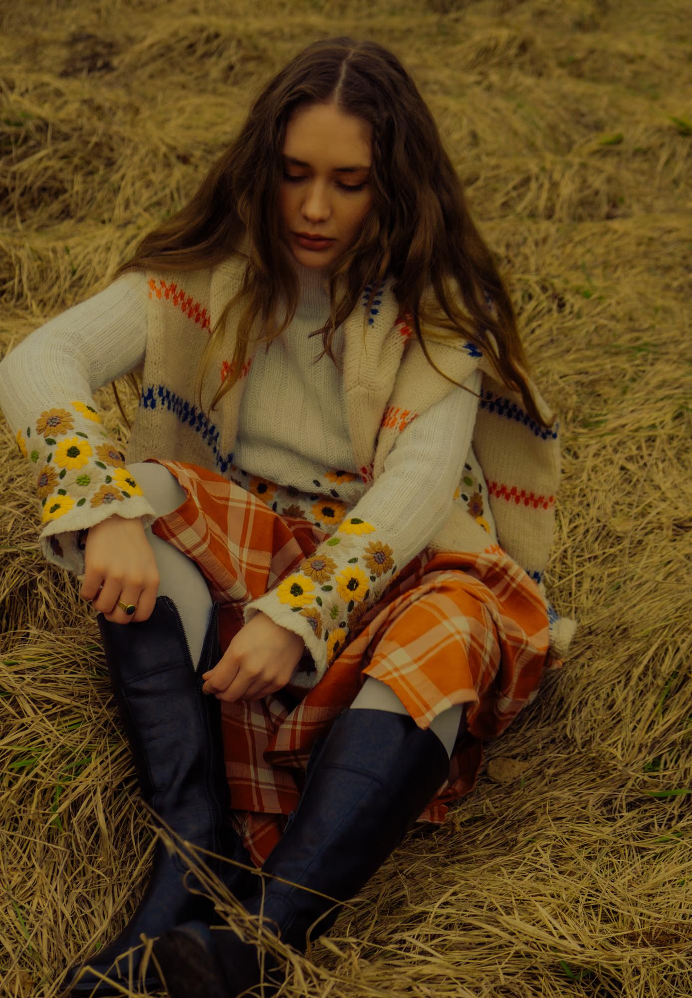 Medium fashion shot of woman in white blouse and orange tartan skirt sitting in dry Carpathian grass, warm autumn light, relaxed ethno editorial pose