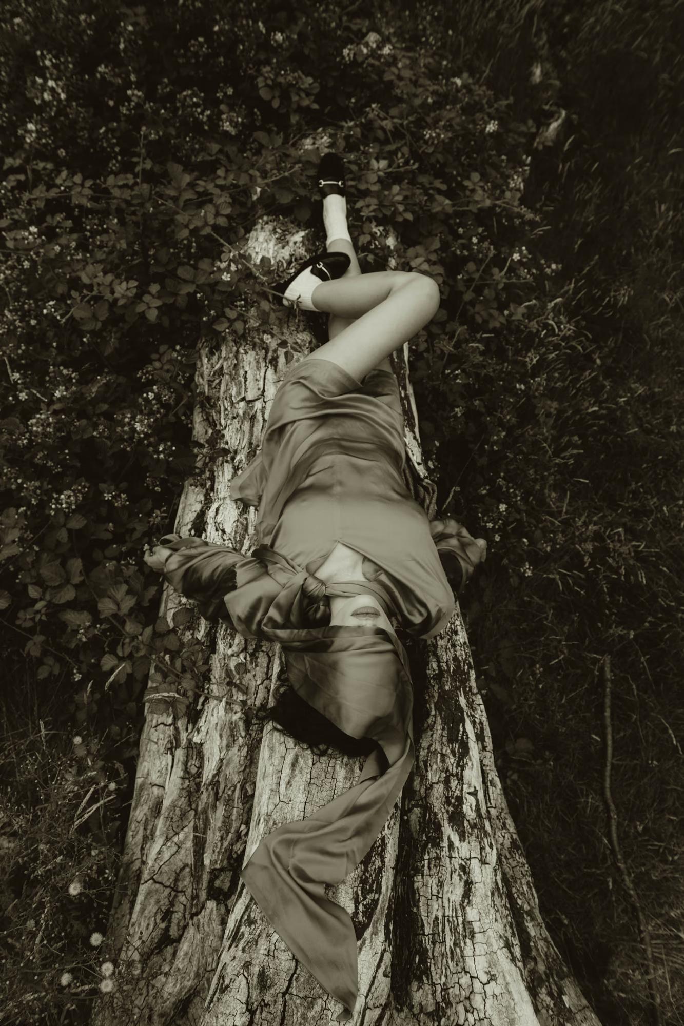Black-and-white photograph shot from above. A model lies draped backwards over a weathered tree stump, her body arched so that her face — barely visible at the bottom — points downward. She wears a fluid satin dress and white socks with black platform shoes. Wild foliage and brambles surround the stump.