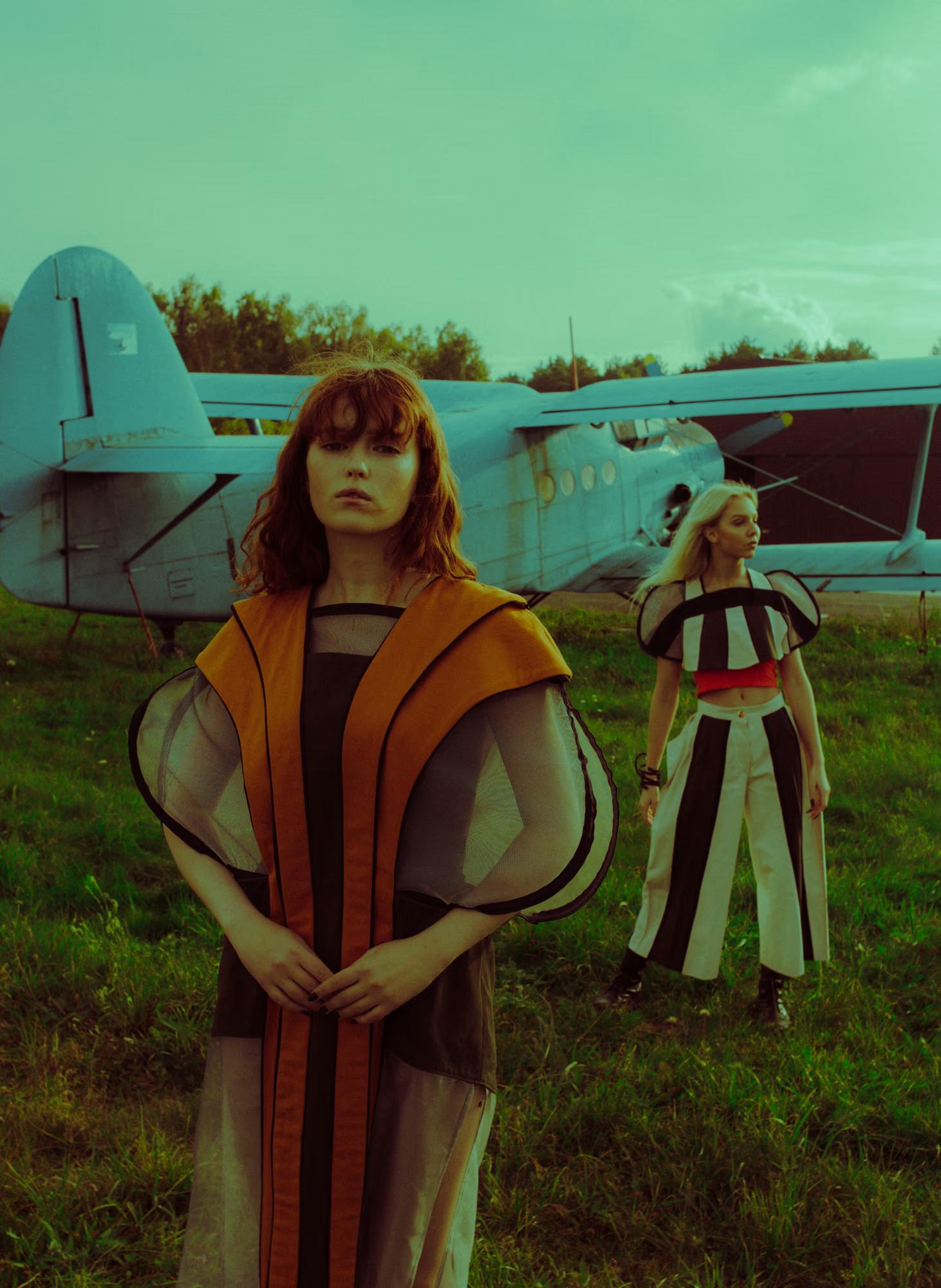 Two models on a grassy airfield with a blue Antonov biplane behind them. The foreground model faces the camera wearing a sheer amber-striped hooded coat over a dark dress. The second model, slightly behind, wears a bold black-and-white striped crop top and wide-leg trousers with a red waistband. Teal colour grade.