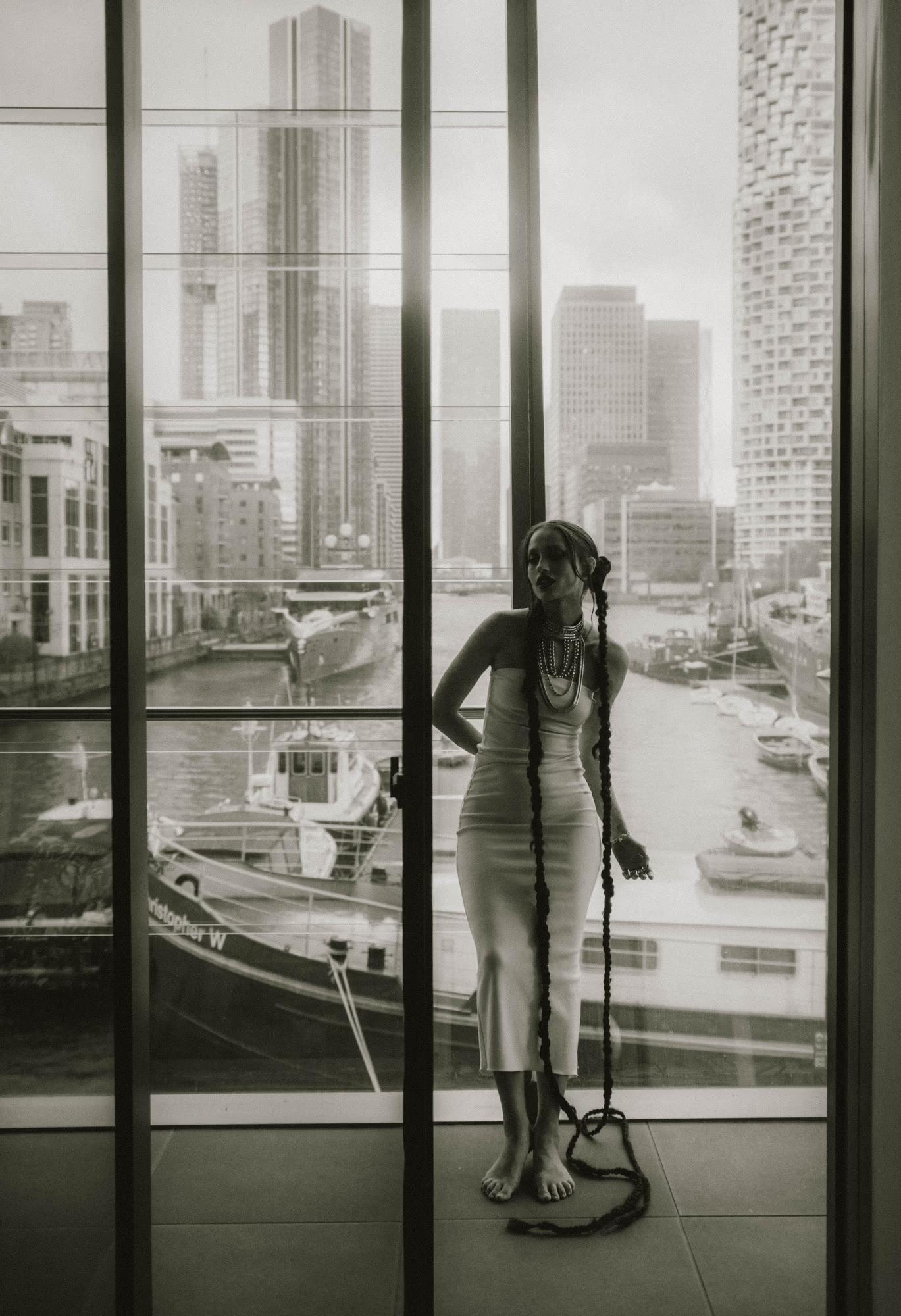  Full body black and white shot of woman standing in front of large industrial windows or glass facade, Canary Wharf London