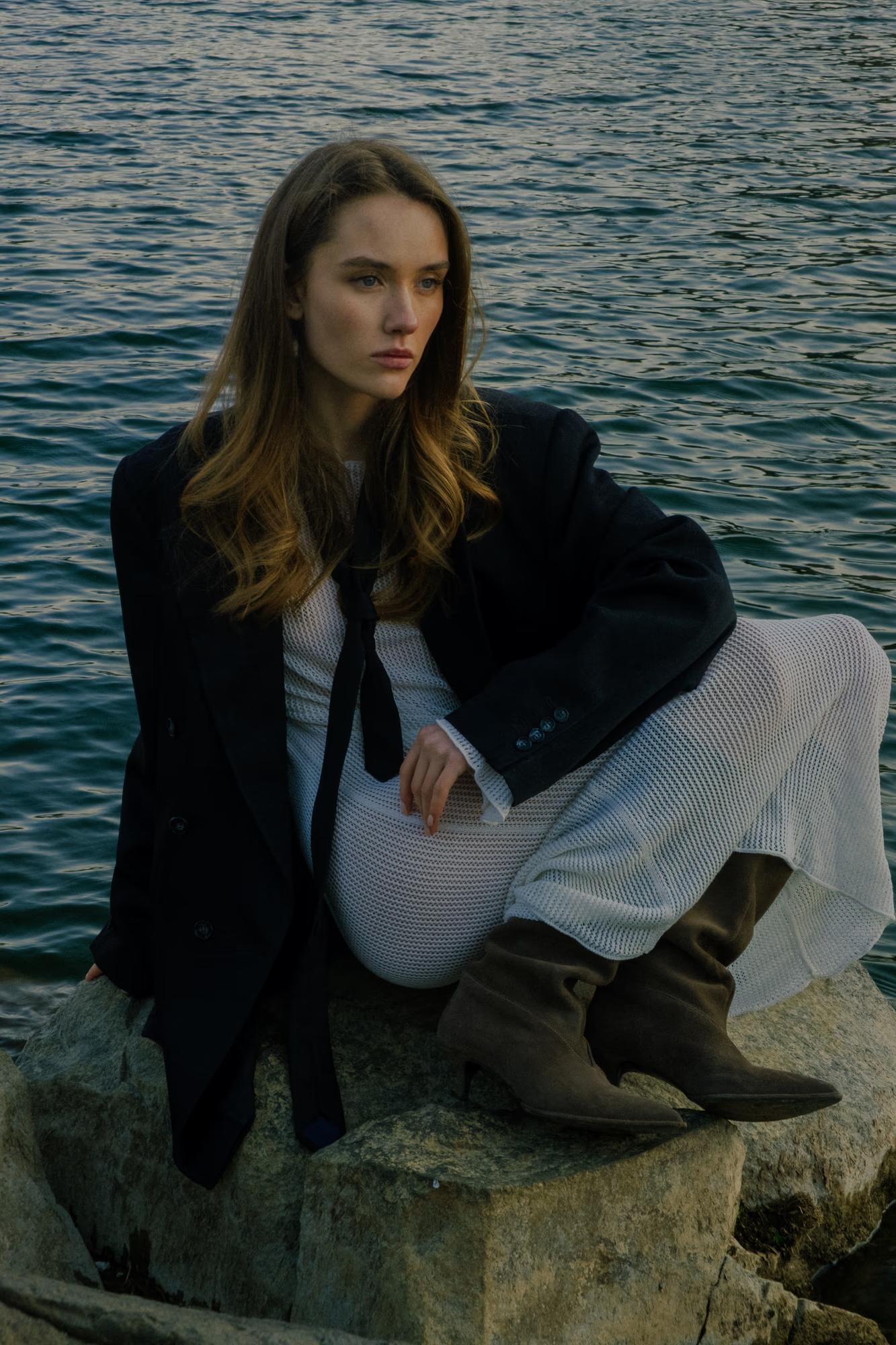 Cinematic pose: The model sits on natural rocks next to the lake, looking contemplative, in black and white fashion.