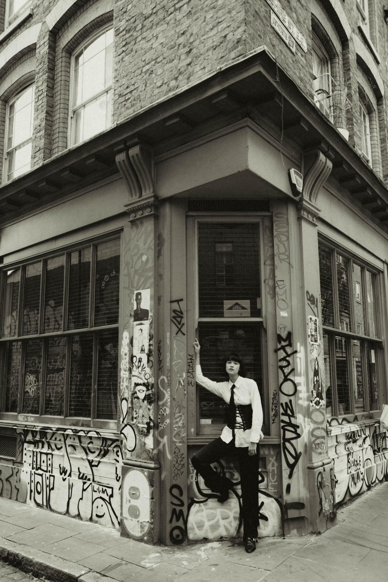 Black and white full body fashion shot of woman in tailored outfit on Shoreditch London street corner with classic brick building, urban editorial