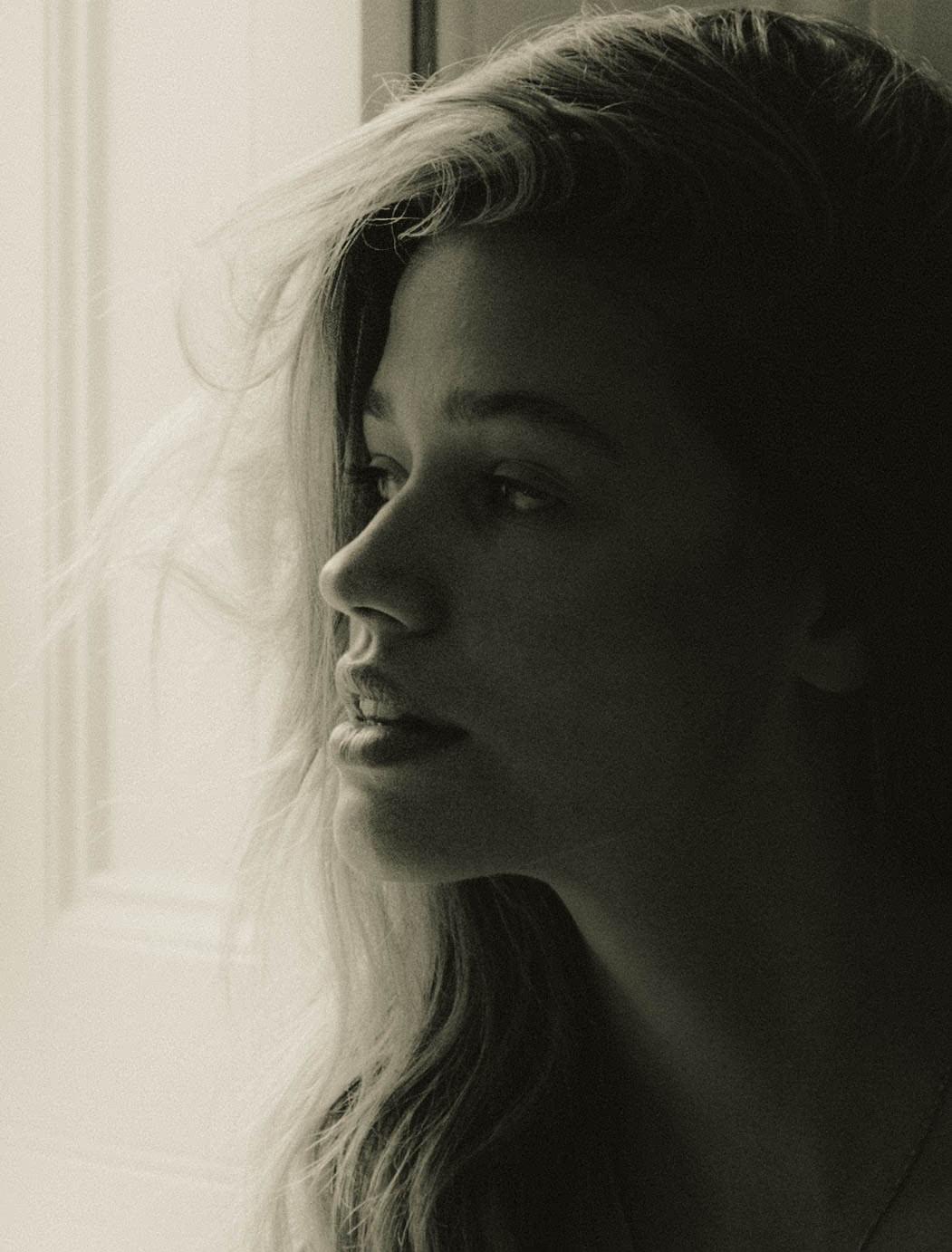Black-and-white close-up side profile portrait of a model with long wavy hair, lit from a bright window to her left. The right half of her face falls into deep shadow, creating a stark, painterly contrast. Her gaze is directed slightly forward, lips slightly parted.