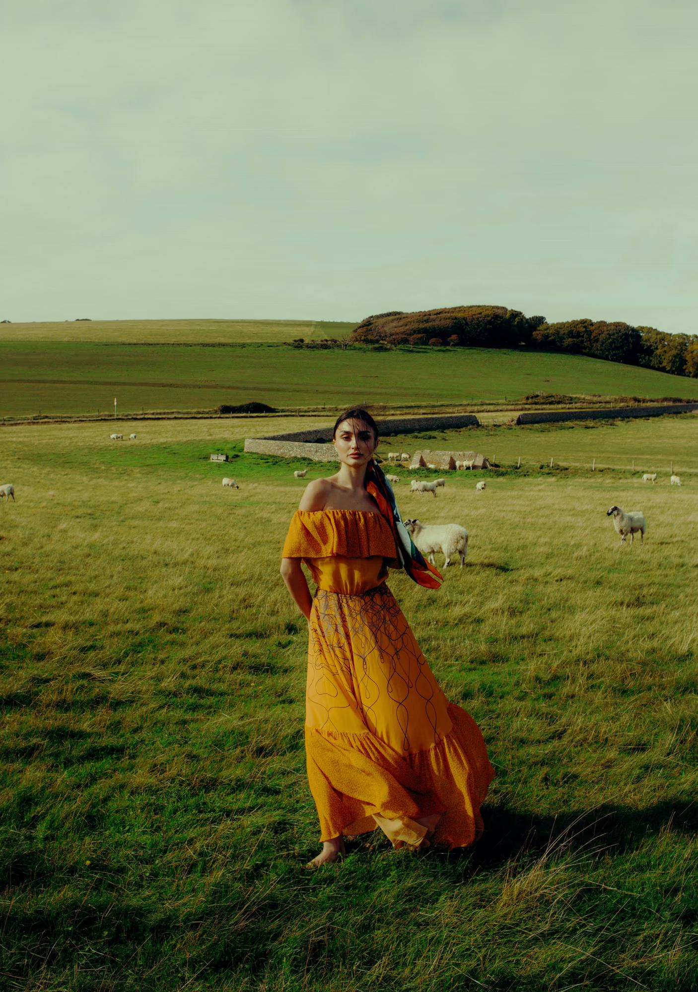 Full body fashion campaign shot of woman in vibrant yellow-orange designer dress in green coastal meadow, Eastbourne cliffs in background, luxury fashion editorial