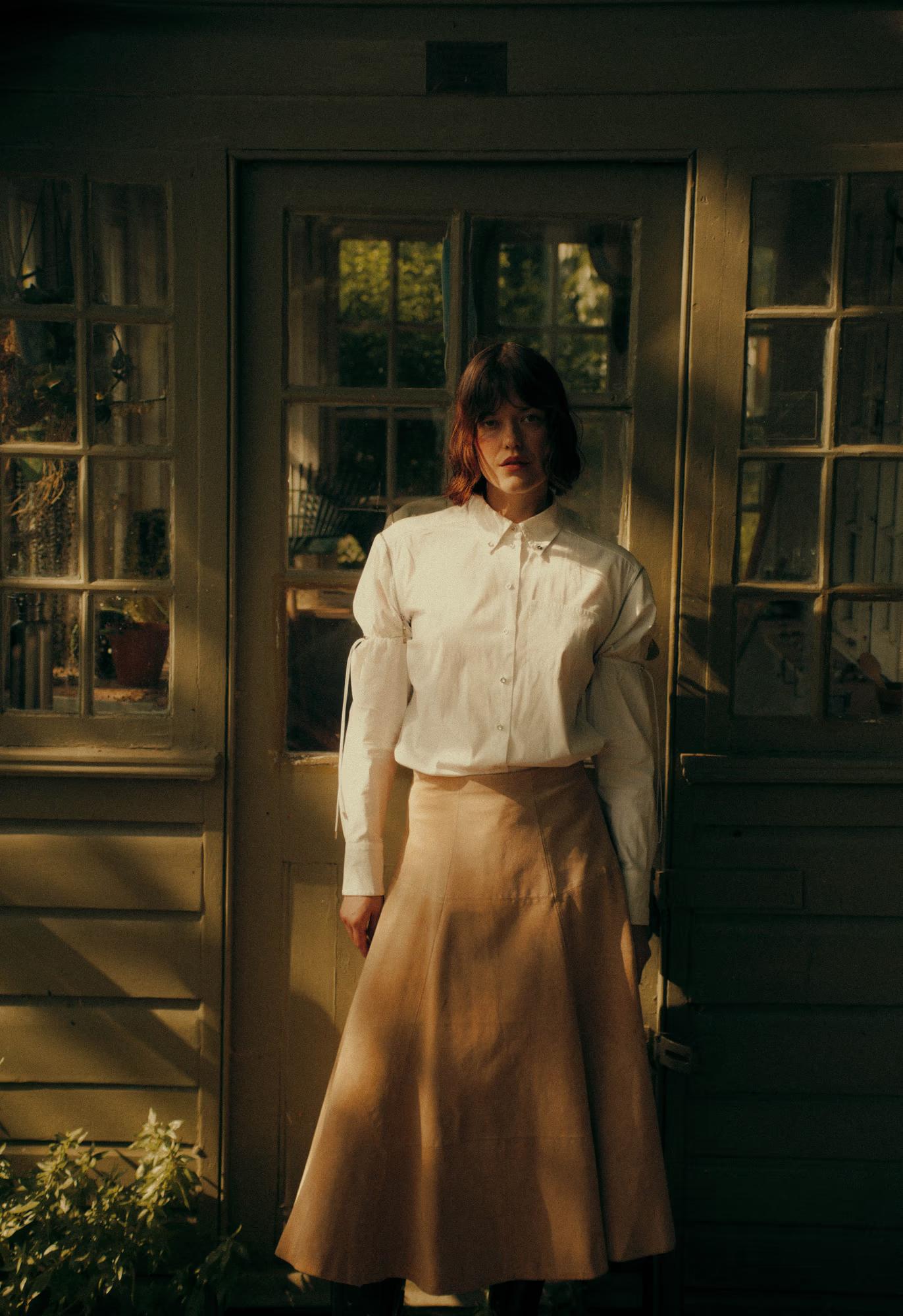 Full-length portrait of a model standing in a doorway of a weathered wooden garden house. She wears a white structured long-sleeve shirt tucked into a flared sand-coloured midi skirt. Warm golden light casts shadows across the peeling doorframe and old window panes beside her.