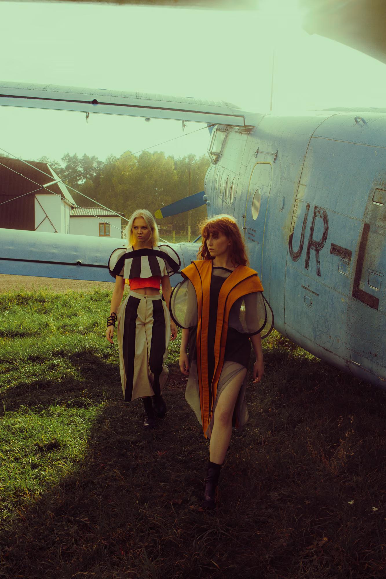 Two models walking away from a pale blue Antonov biplane on an airfield. The blonde model leads in a black-and-white graphic wide-leg outfit; the redheaded model follows in an amber-panelled sheer coat. The registration "UR-L…" is visible on the aircraft fuselage. Overcast, teal-tinted light.