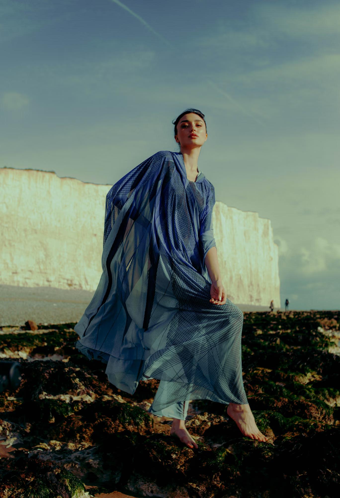 Full body fashion campaign shot of woman in dramatic blue designer cape on rocky Eastbourne coastline, moody overcast sky, luxury editorial