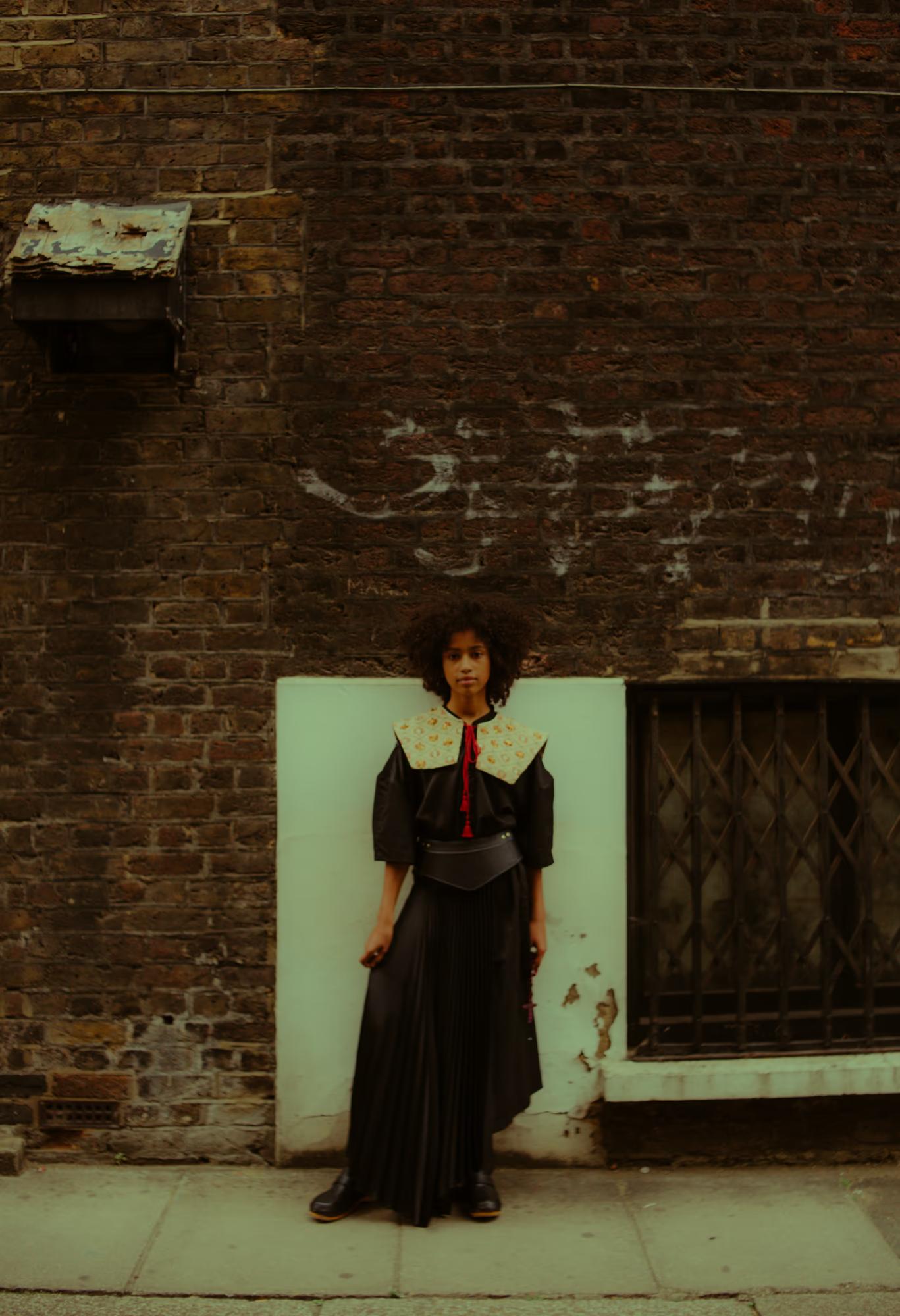 Full-length portrait of a model standing against a dark, weathered brick wall in a London back alley. She wears a black blouse with a wide embroidered folk collar, a red tassel tie, a leather belt, and a long black pleated skirt with black clogs. Her expression is calm and direct.