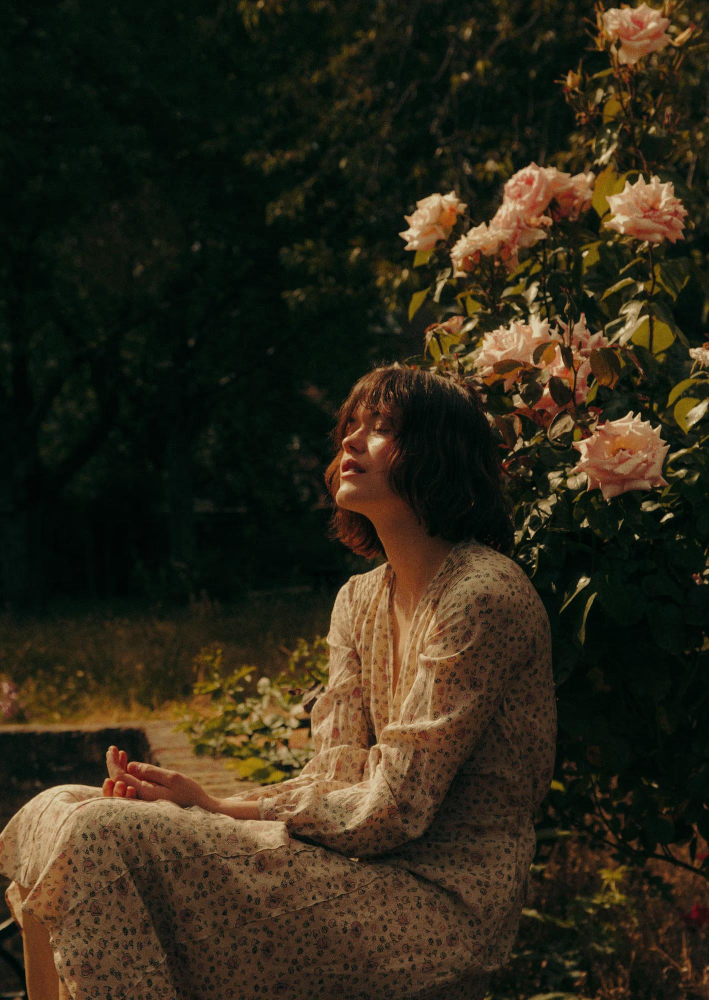 A model sits on a stone ledge in dappled sunlight beside a large bush of soft pink roses, her face tilted upward with eyes closed. She wears a flowing long-sleeve floral dress in cream and pale tones. The dark background of trees contrasts with the warm light falling on her.