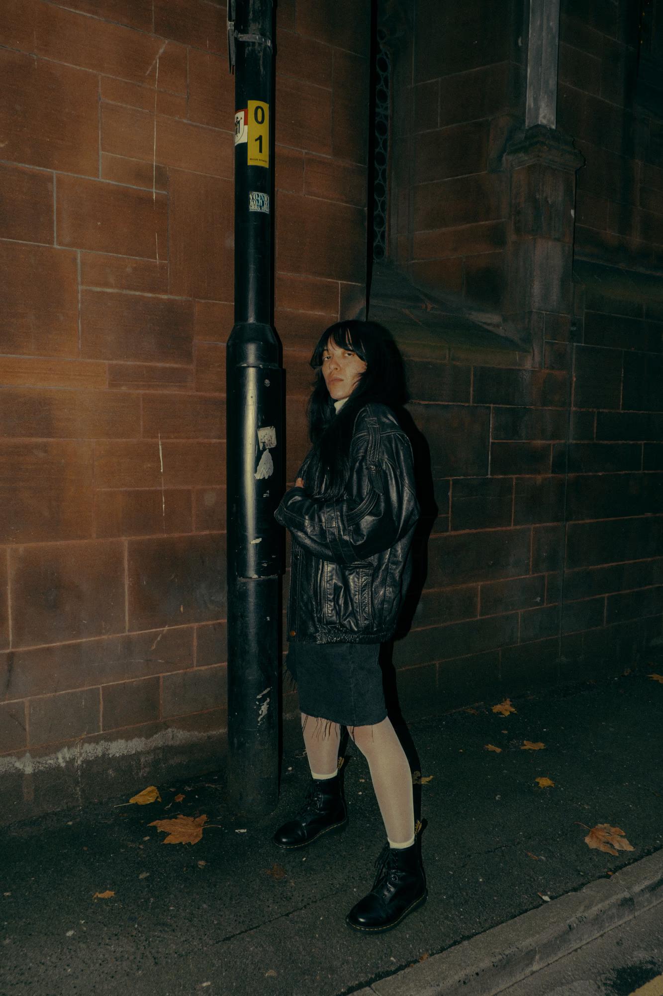 Full body fashion shot of person in dark patterned coat walking on Manchester street with brick building facade, moody cinematic urban editorial