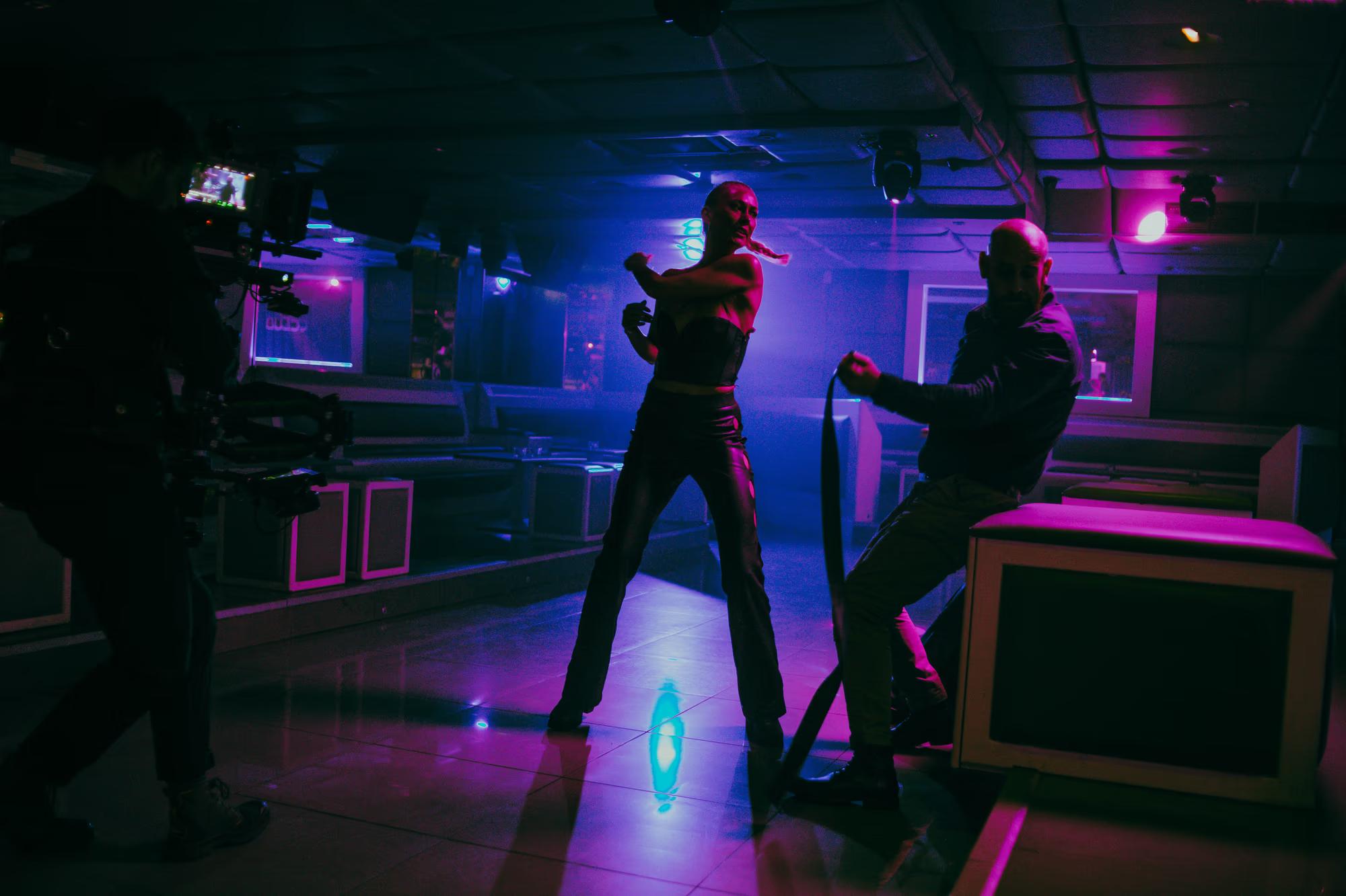 Stylized action shot with neon blue and pink lighting, showing the crew filming a modern fight sequence.