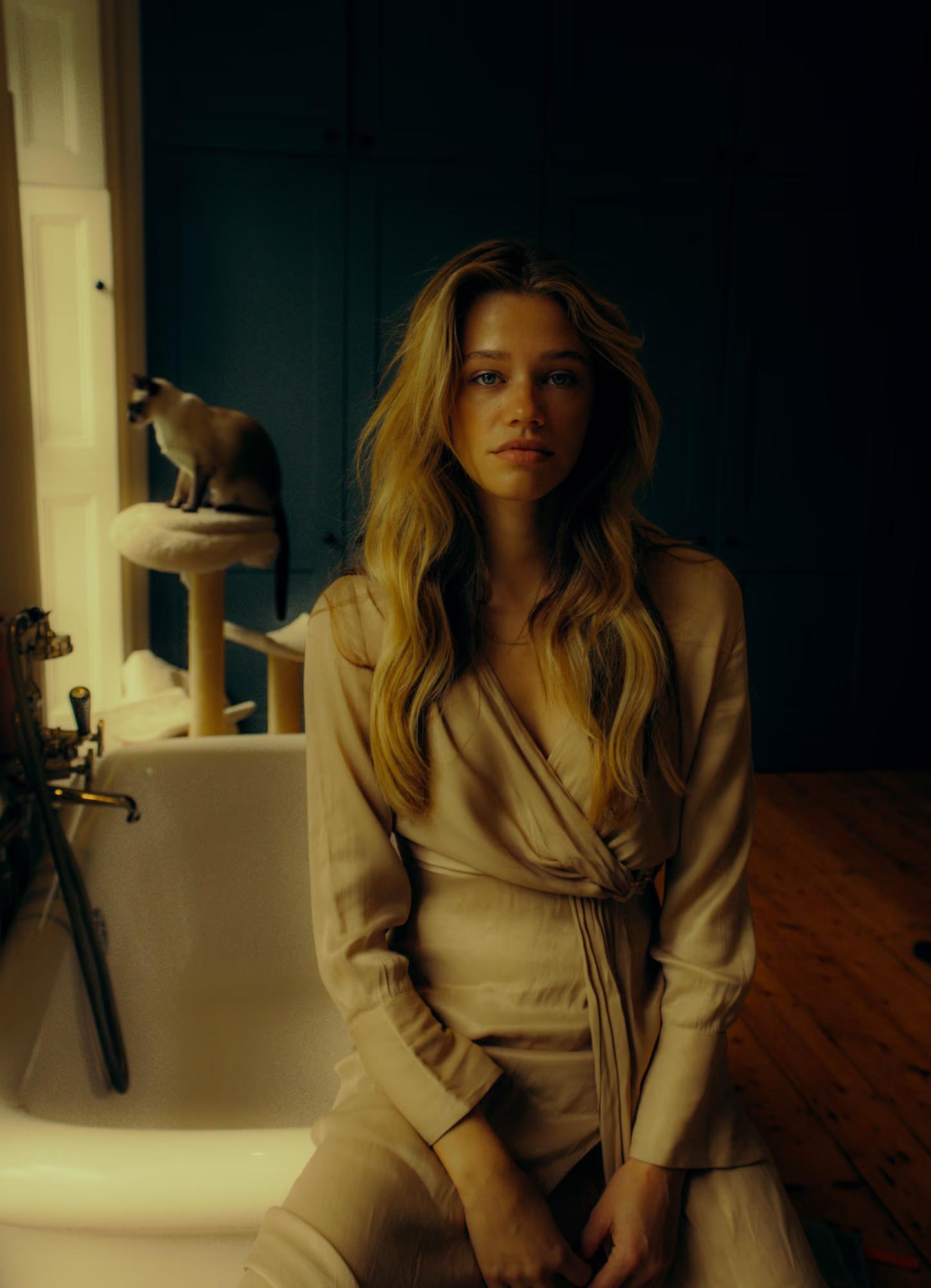 A model sits on the edge of a freestanding bathtub in a room with dark teal walls and worn wooden floorboards. She wears a long-sleeve sandy-coloured wrap dress and looks directly at the camera. Behind her, a Siamese cat perches on a cat tree by the window, bathed in natural light.