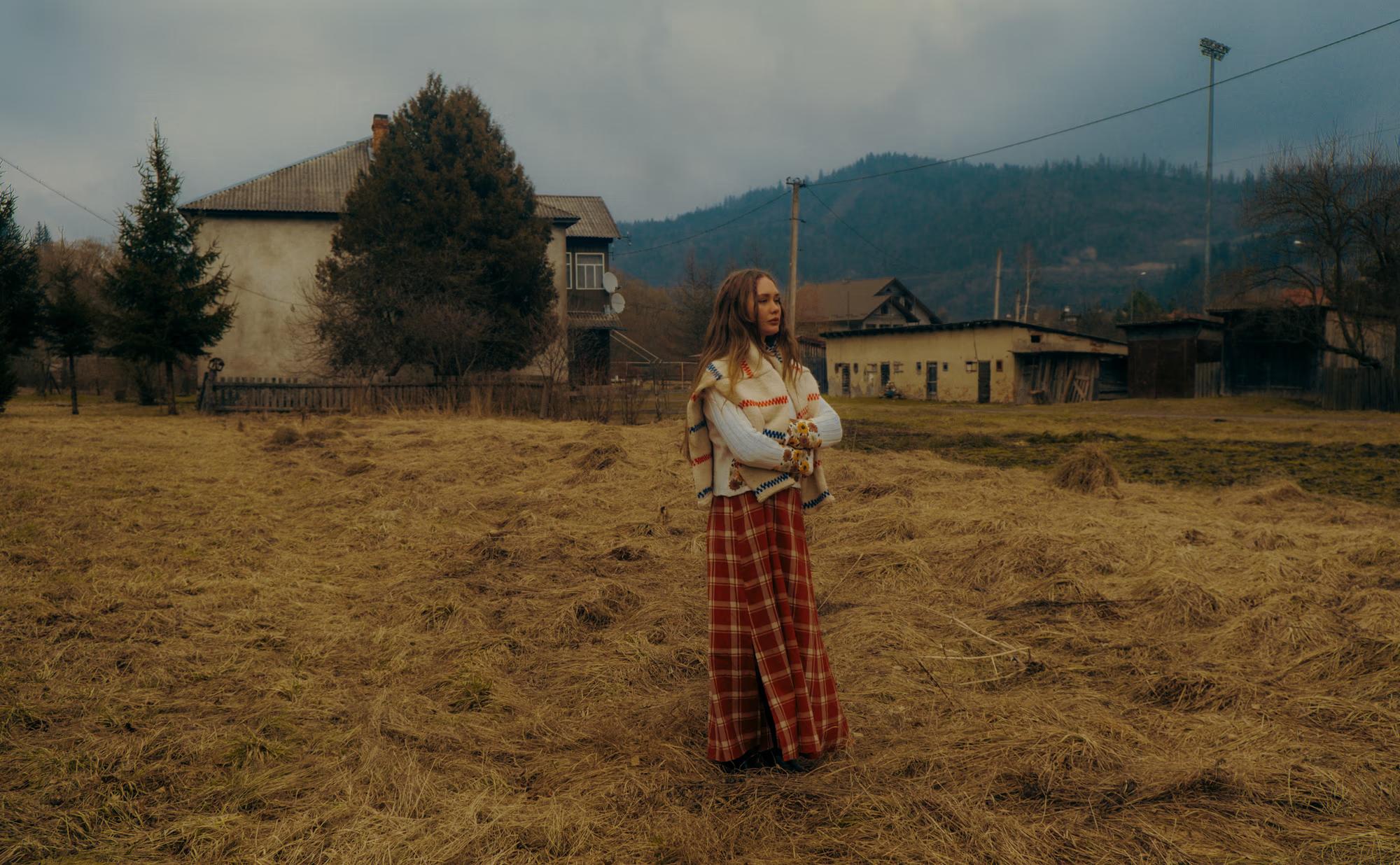 Wide fashion shot of woman in white blouse and red tartan skirt walking through dry golden Carpathian meadow with mountain village and peaks in background, Ukrainian ethno editorial