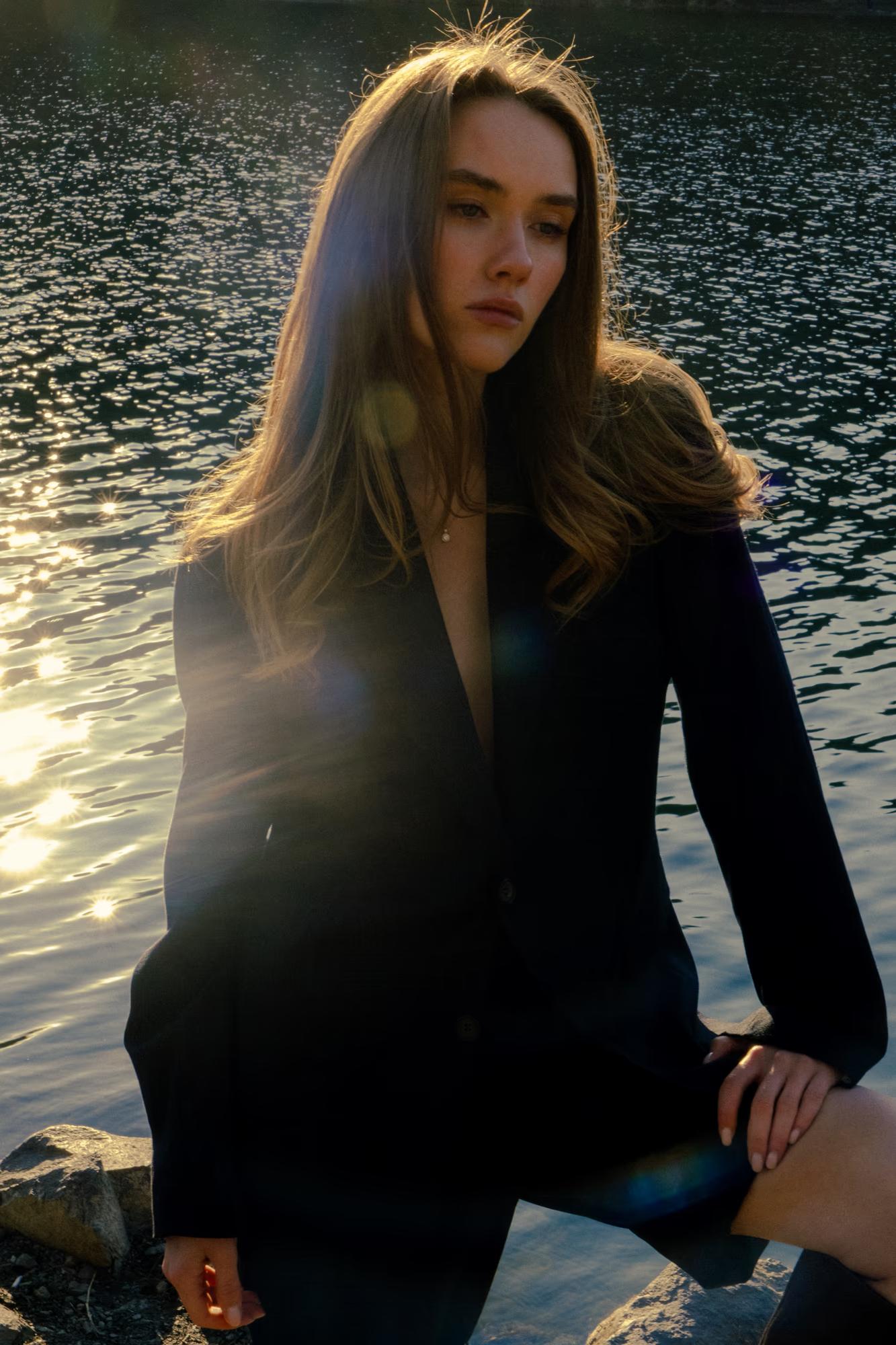 Direct, confident pose of the model standing near the rocky lake shore, wearing a structured blazer and an airy dress.