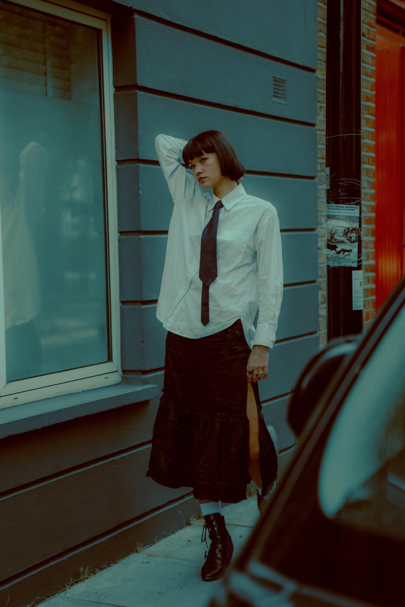 Full body fashion campaign shot of woman in androgynous tailored outfit with red skirt standing against teal wall, bold color contrast, Shoreditch London street editorial