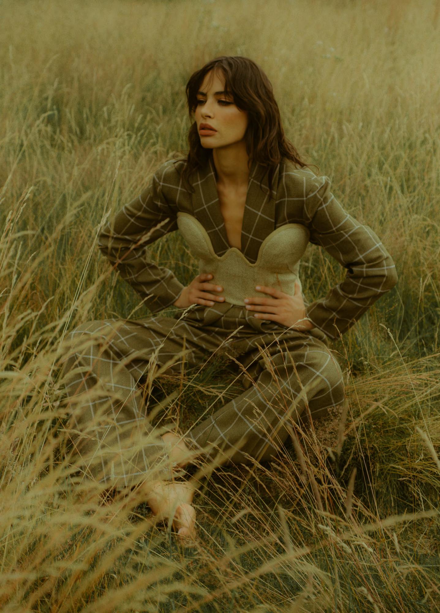 A model sits barefoot amid tall golden grass, wearing an open-front olive green windowpane check blazer and matching wide-leg trousers. Underneath she wears a structured nude strapless corset with a scalloped top edge, which she holds with both hands. She gazes softly downward.
