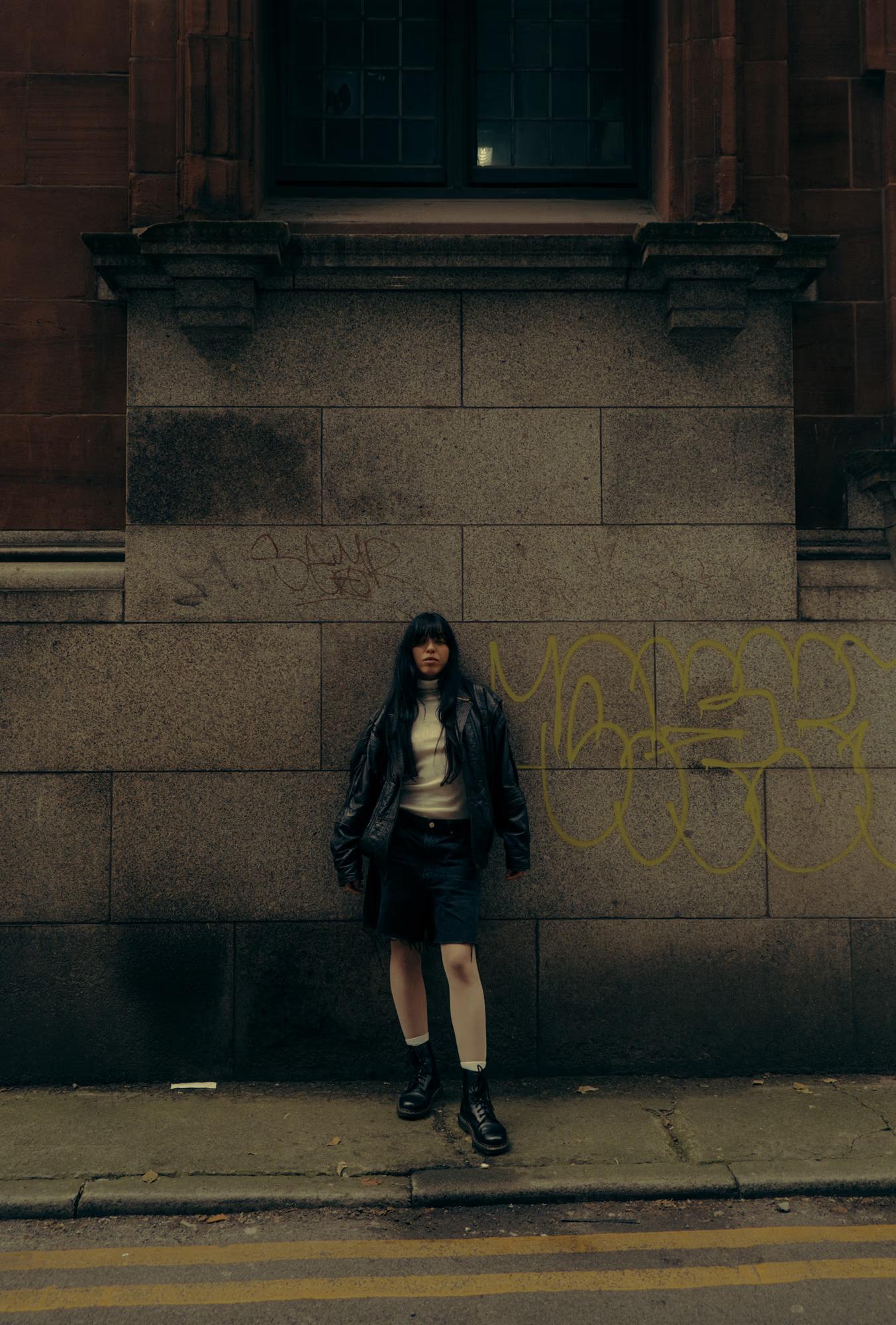 Full body fashion shot of person in dark outfit standing alone on wide Manchester street pavement with brick wall backdrop, moody cinematic editorial